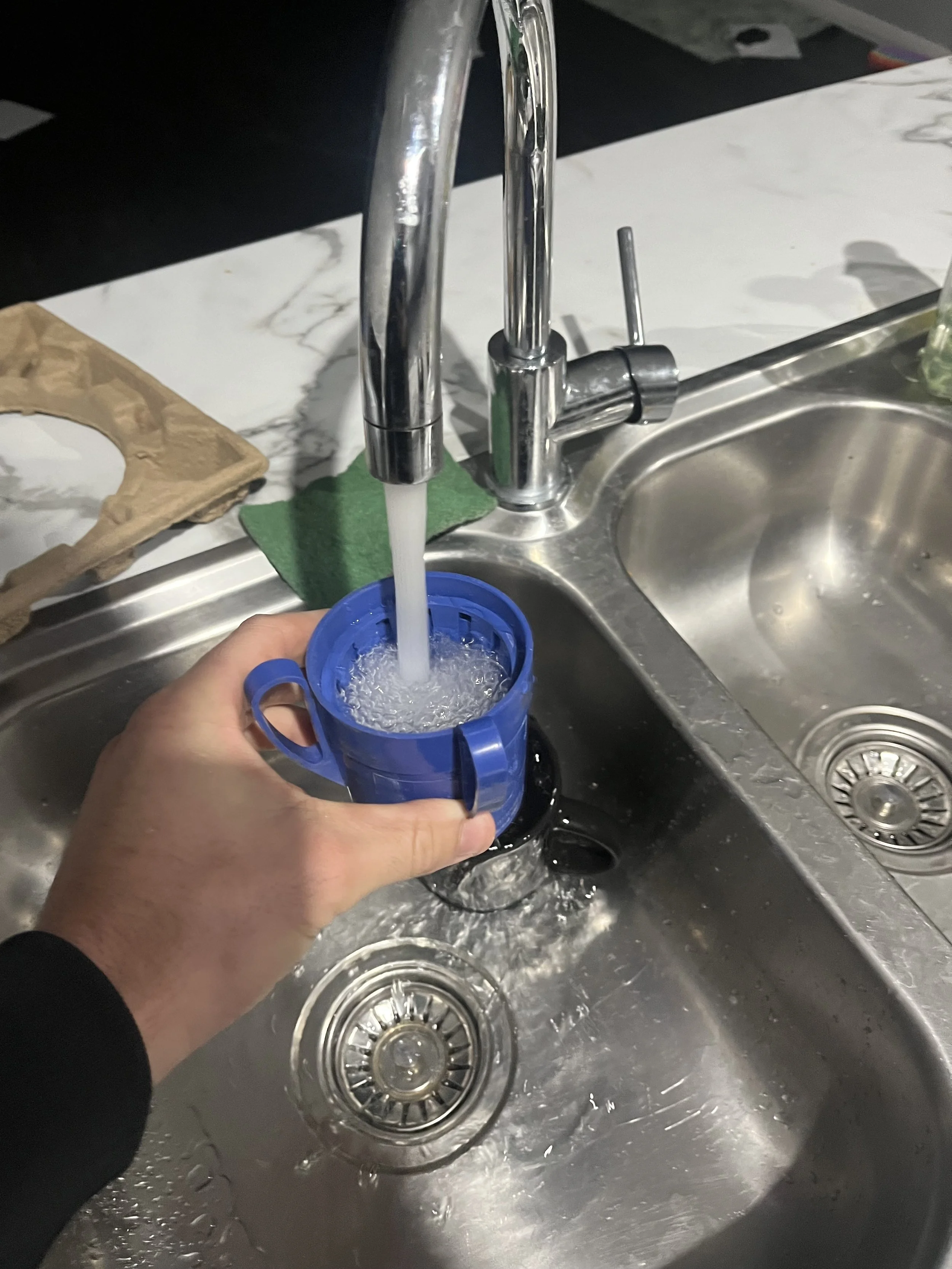 Hand holding a blue mold under running water in a stainless steel kitchen sink.