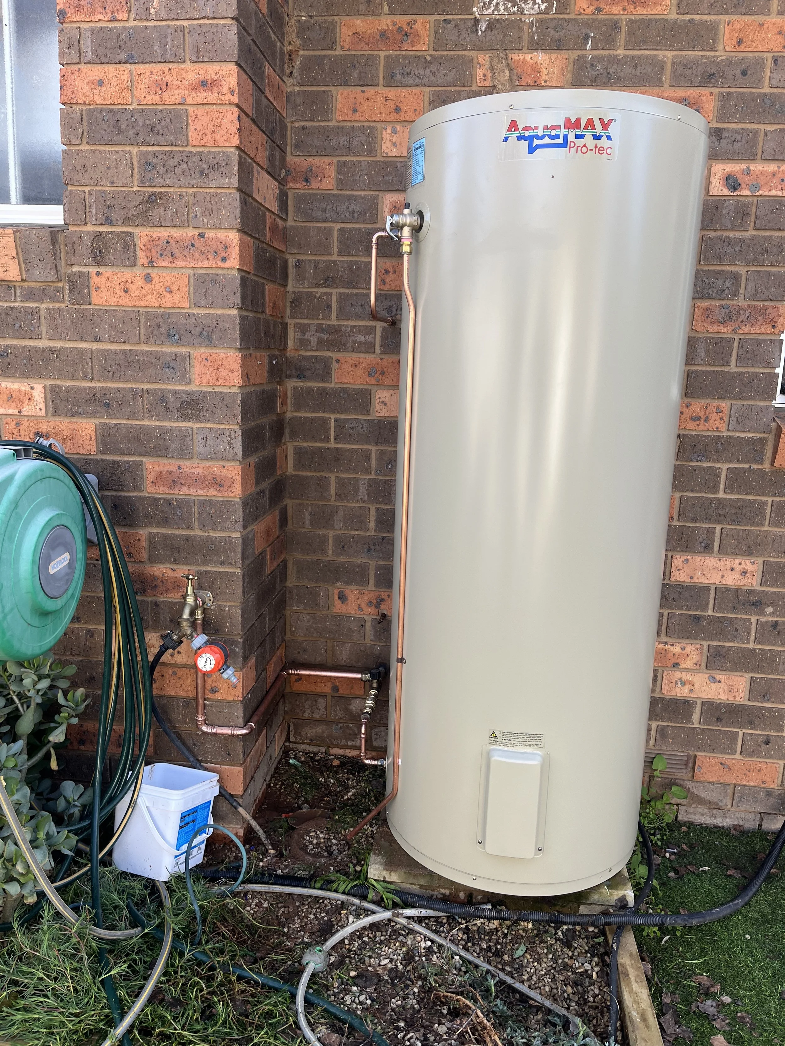 Outside view of a water heater tank next to a brick wall, with pipes and hoses connected, and a green garden hose reel on the left.