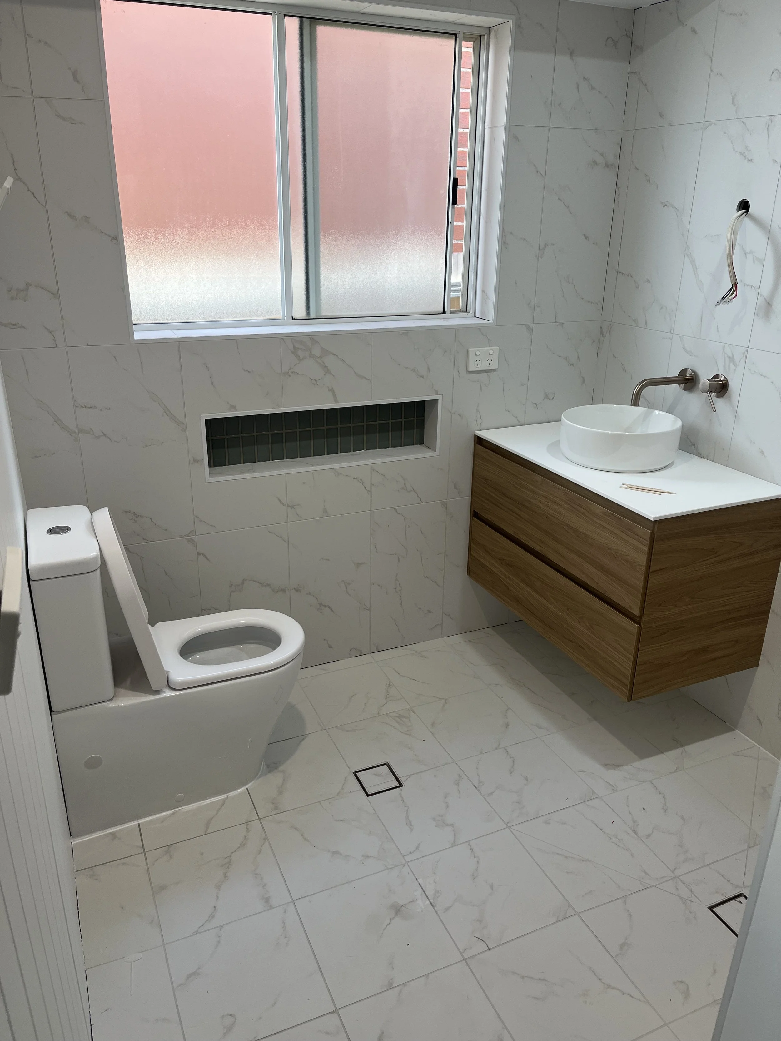 Empty modern bathroom with white marble tiles, a frosted window, a white toilet, a floating wooden vanity with a round white vessel sink, and a wall-mounted faucet.