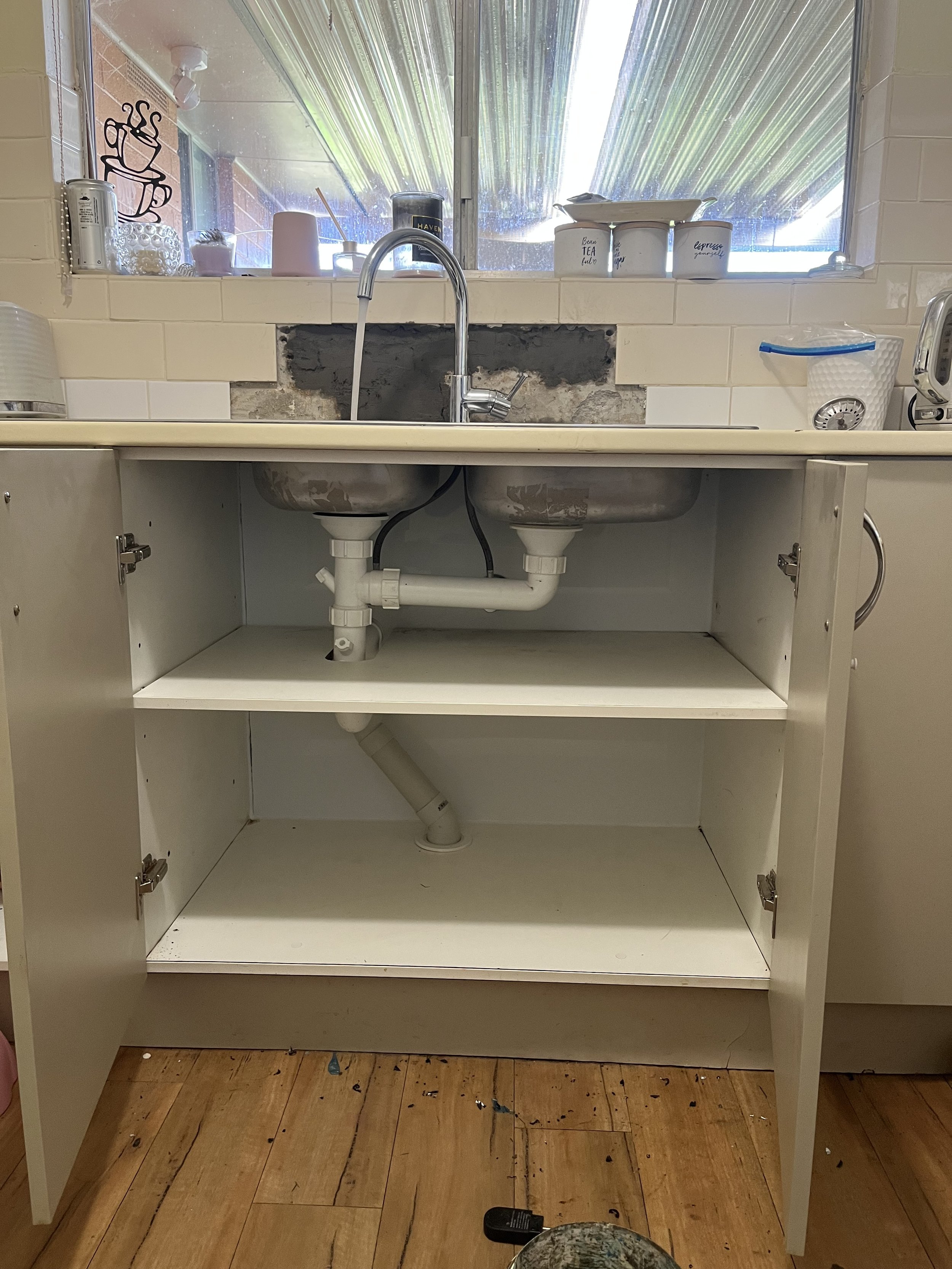 Open kitchen cabinet under a sink showing plumbing pipes inside the cabinet and a white countertop with a double sink and a faucet above.