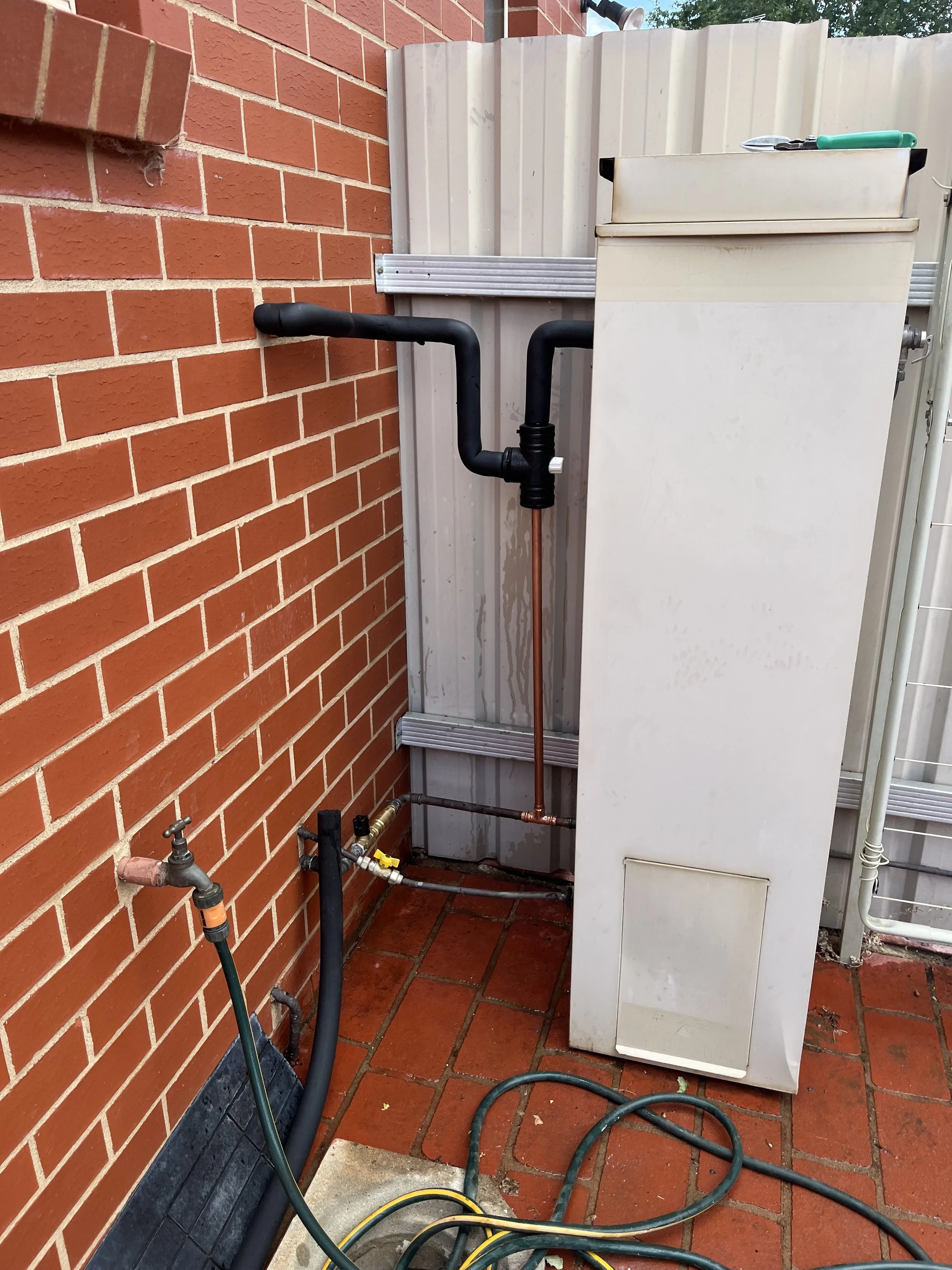Outdoor water heater with black pipes, a garden hose, and a faucet next to a brick wall and a metal fence.