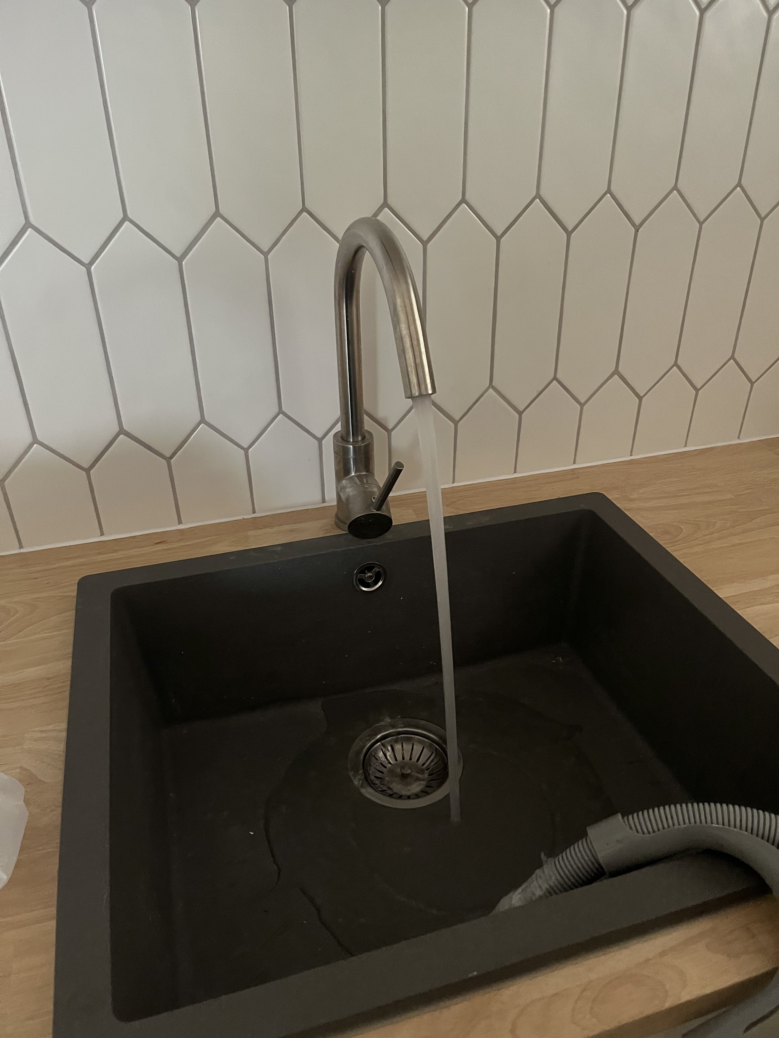 A black kitchen sink with a silver faucet, with water running into it, set into a wooden countertop with a white hexagonal tile wall in the background.