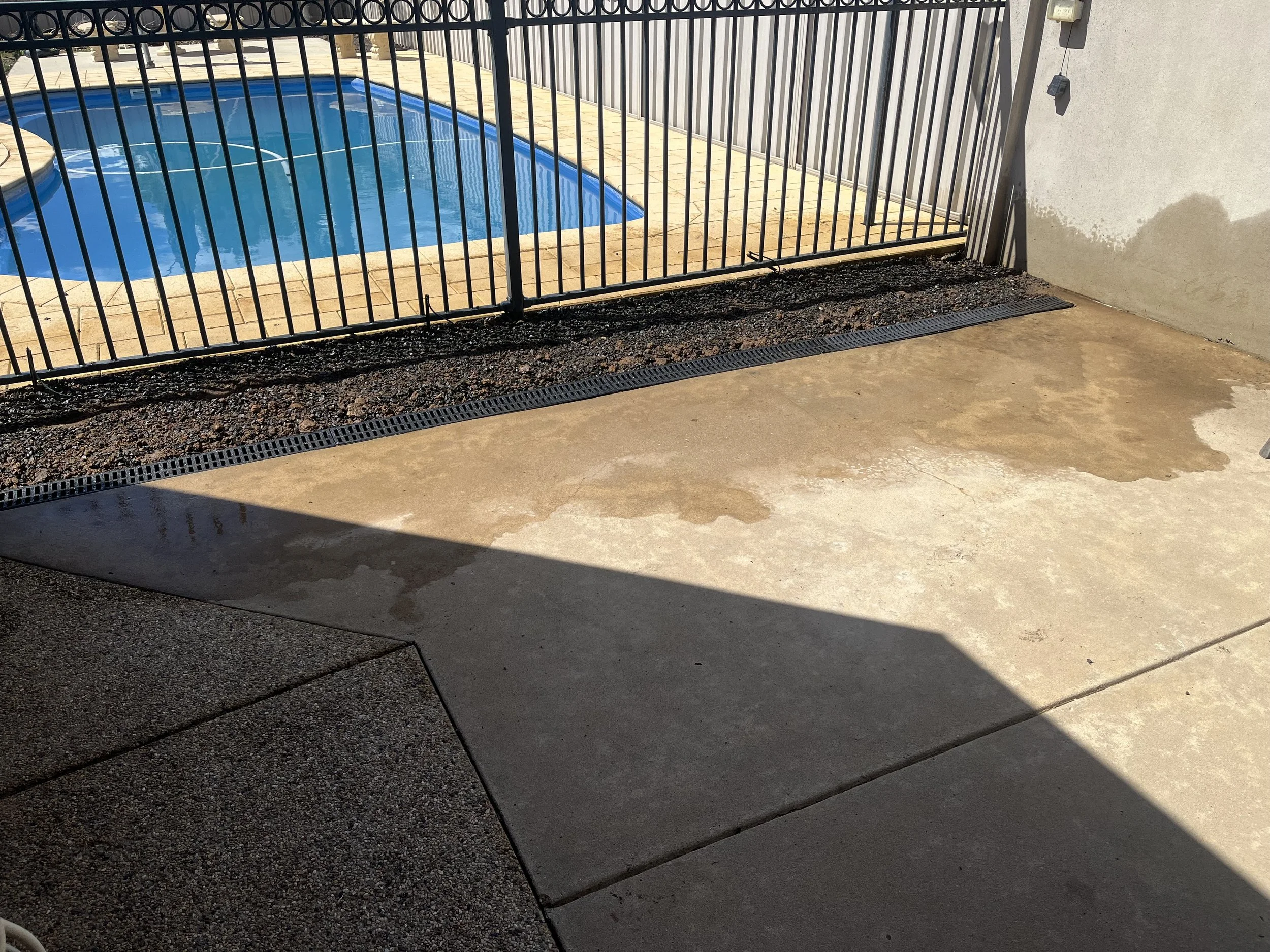 A section of a backyard pool area with a black metal fence, concrete patio, and a shadowed area.