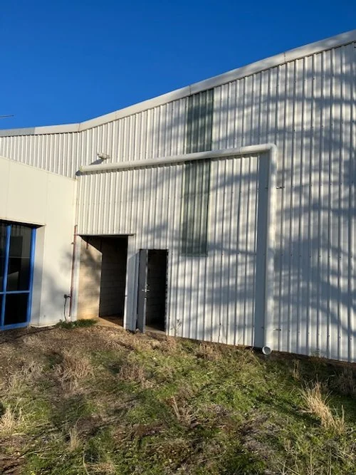 Side of a white industrial or warehouse building with a small opening, a pipe, and shadows cast by nearby trees under a clear blue sky.