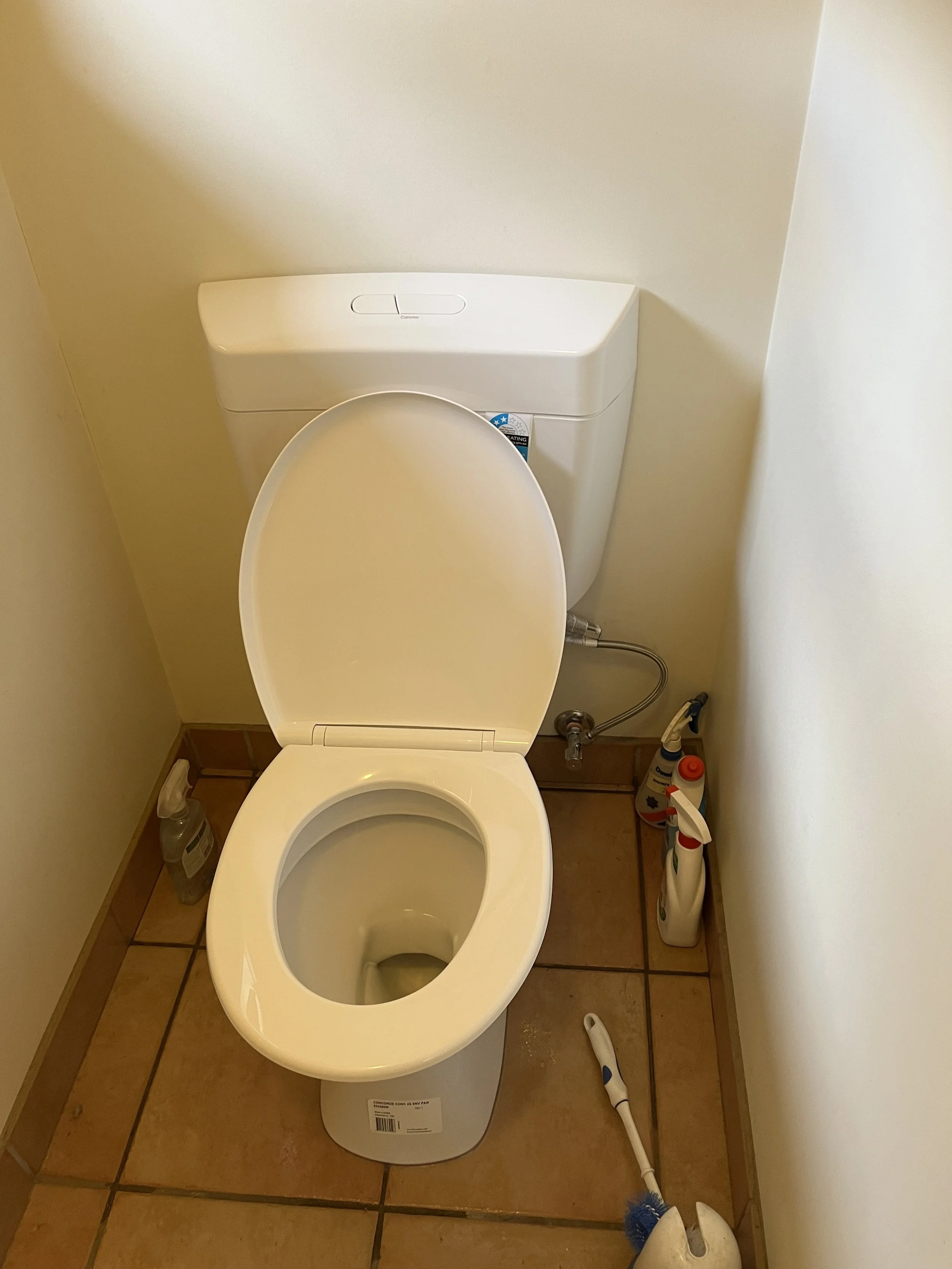 A white toilet with a closed lid in a small bathroom, with cleaning supplies on the floor and a beige tiled floor.