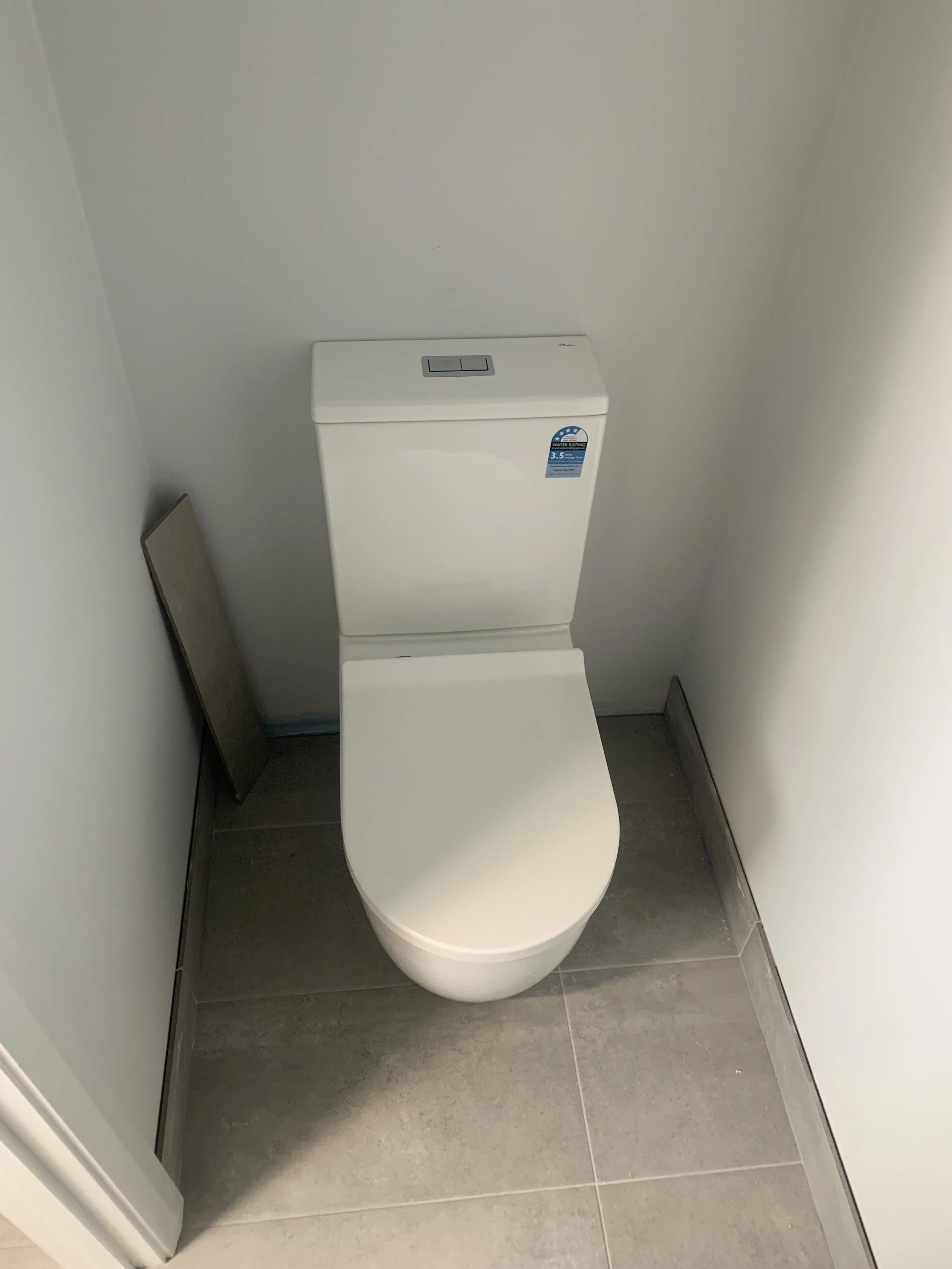 A white toilet with a tank and closed lid, situated in a small bathroom corner with light gray tiled flooring and white walls.