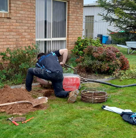 Person digging a hole in the yard with a shovel near a house, using a red portable air compressor.