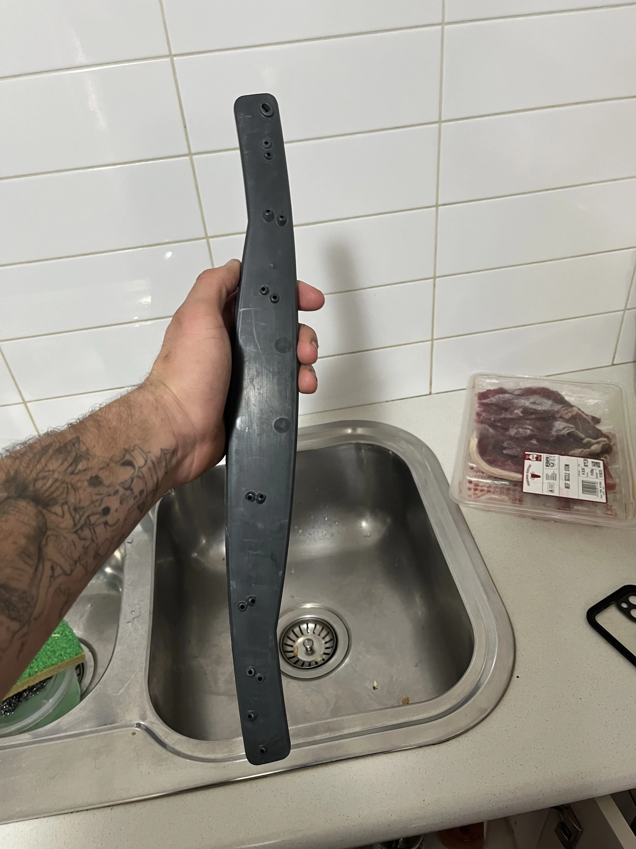 A person's hand holding a black plastic part over a kitchen sink, with a package of raw meat on the countertop to the right.