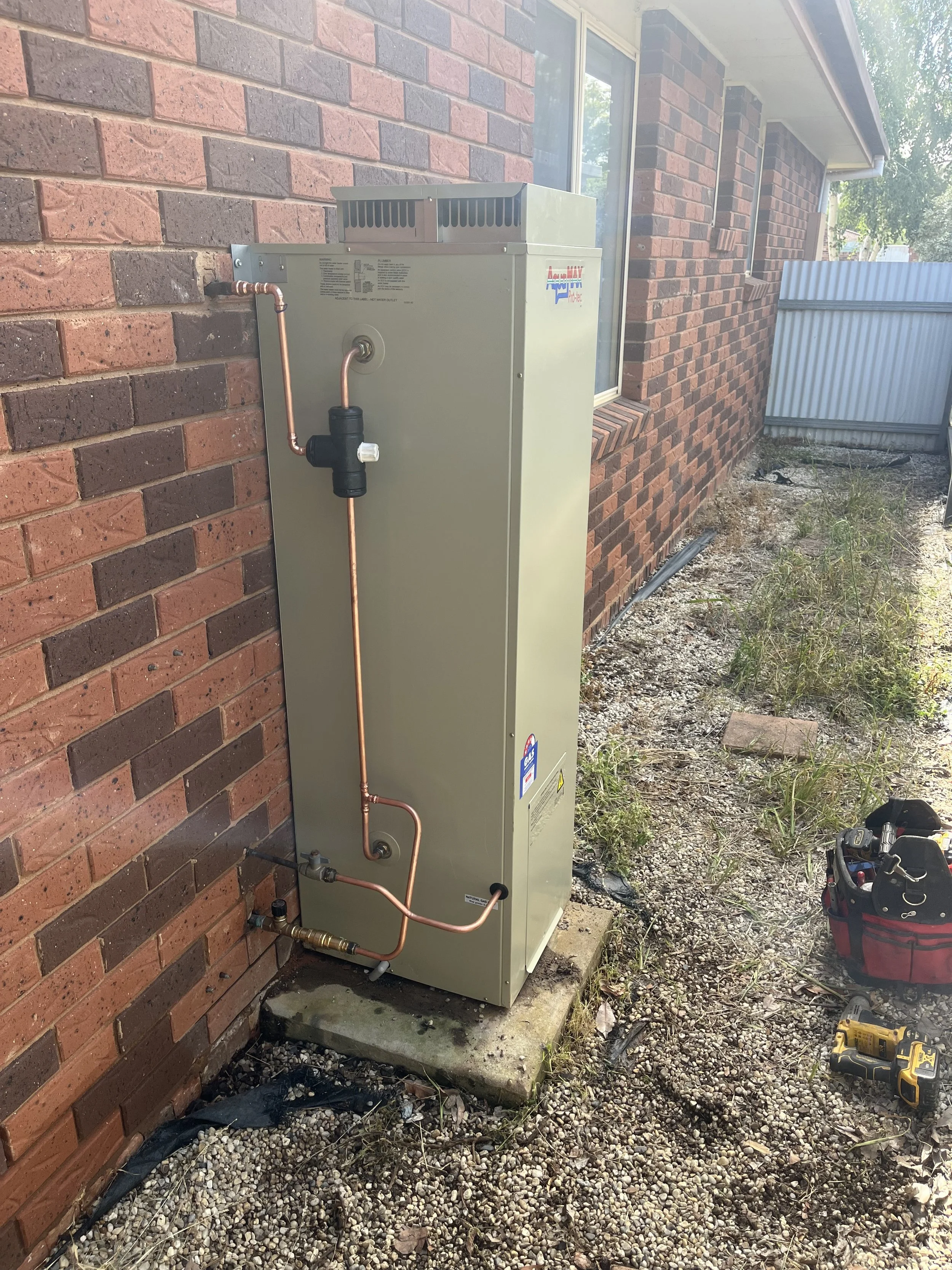 Outdoor hot water heater installation next to a brick house wall, with copper pipes connected to it, on a concrete base, with a toolbox and power drill nearby.