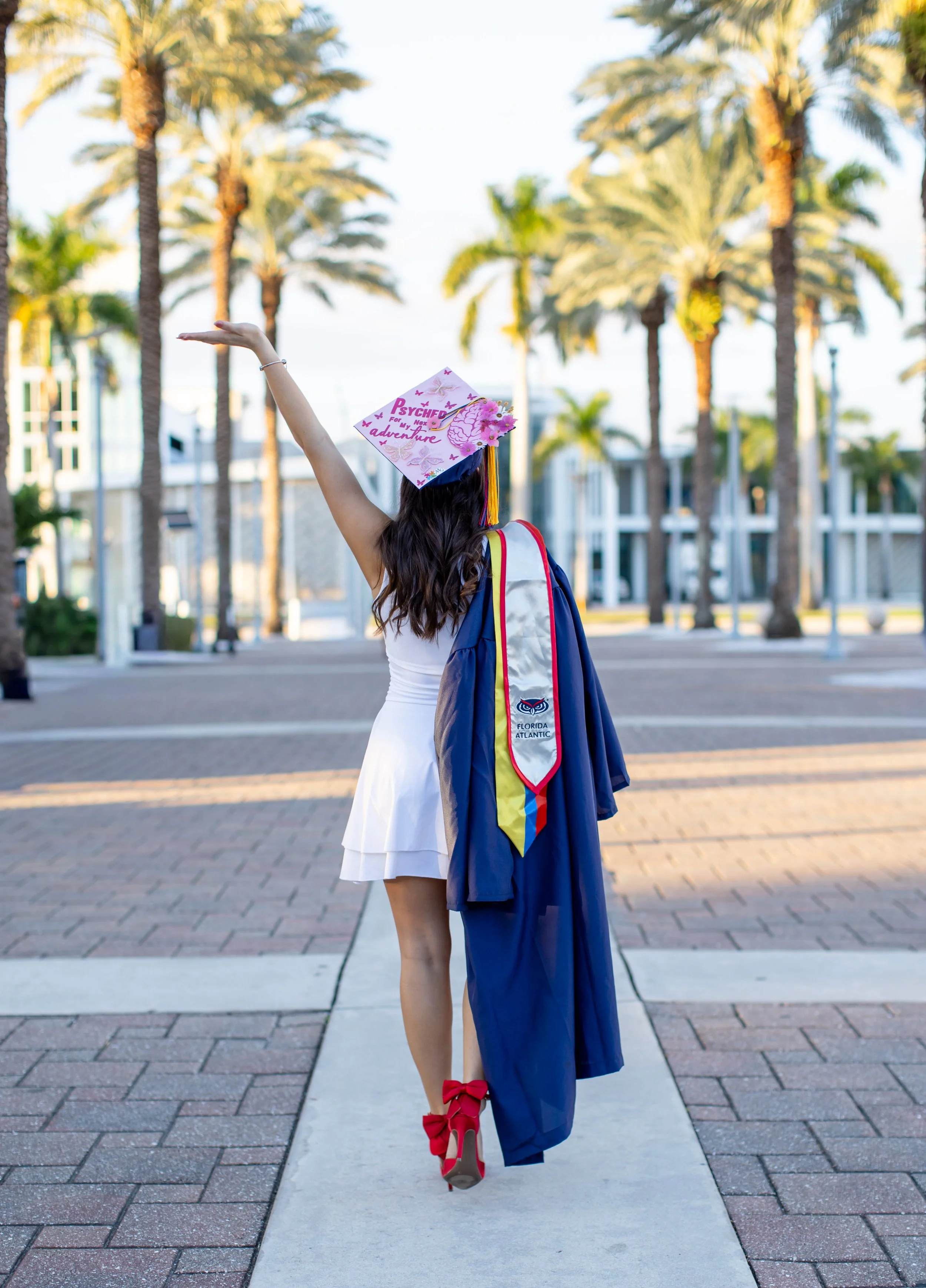 A female graduate in a white dress and red heels with large bows is celebrating her graduation ceremony outside, wearing a graduation cap decorated with pink and purple designs and holding a gown over her arm. She is standing on a sidewalk with palm trees and buildings in the background during dawn or dusk.