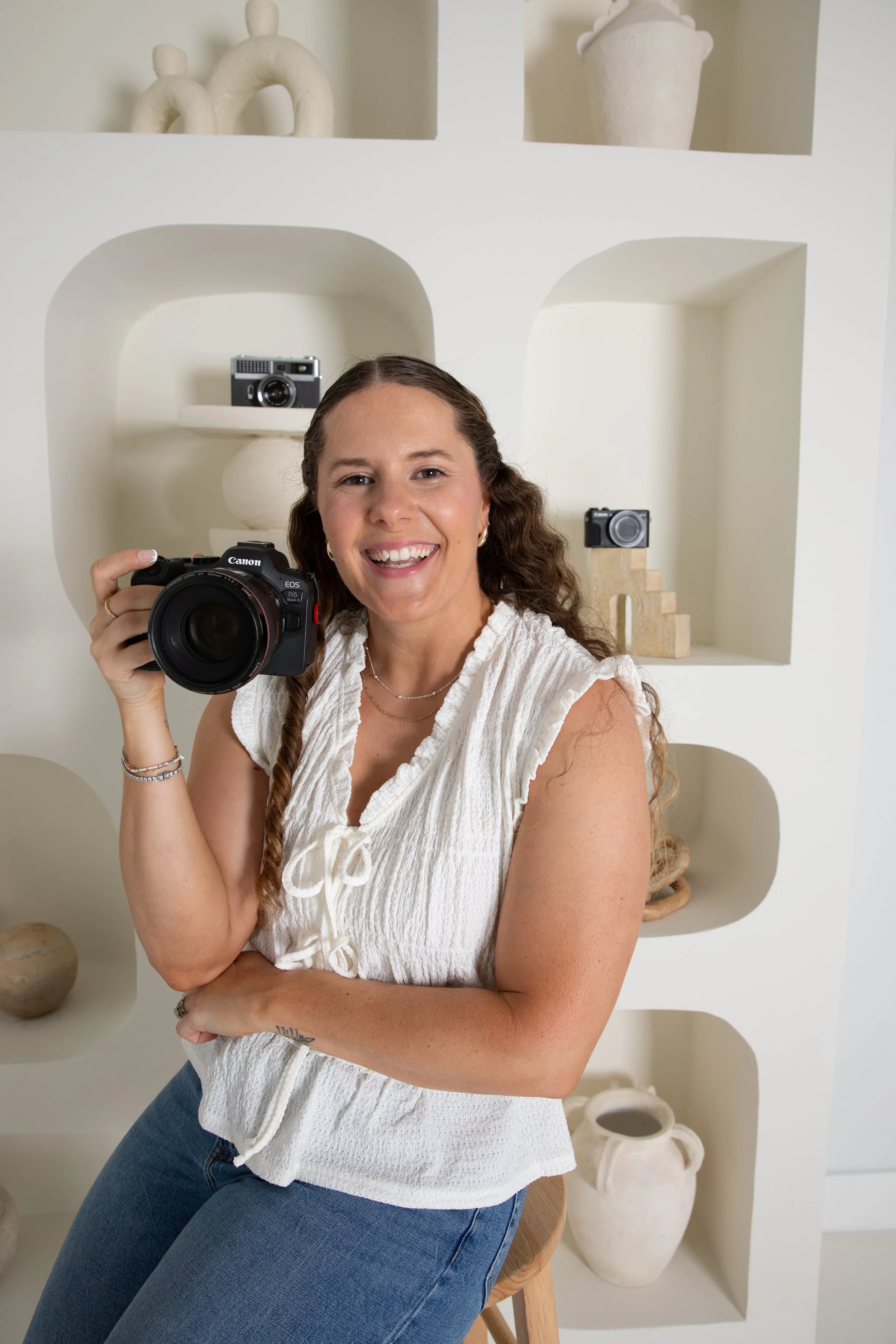 A woman with long curly hair smiling and holding a professional camera, sitting in front of a white gallery wall with decorative ceramics and cameras displayed.