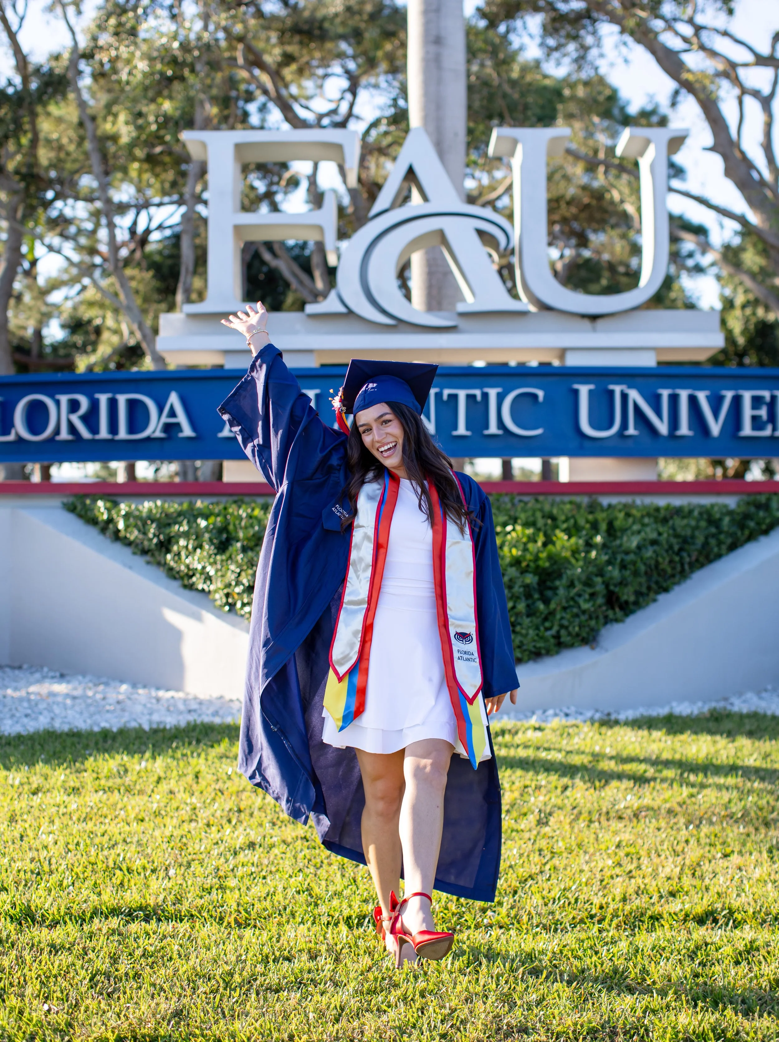 A young woman in a graduation cap and gown celebrating outside Florida Atlantic University with a large FAU sign in the background.