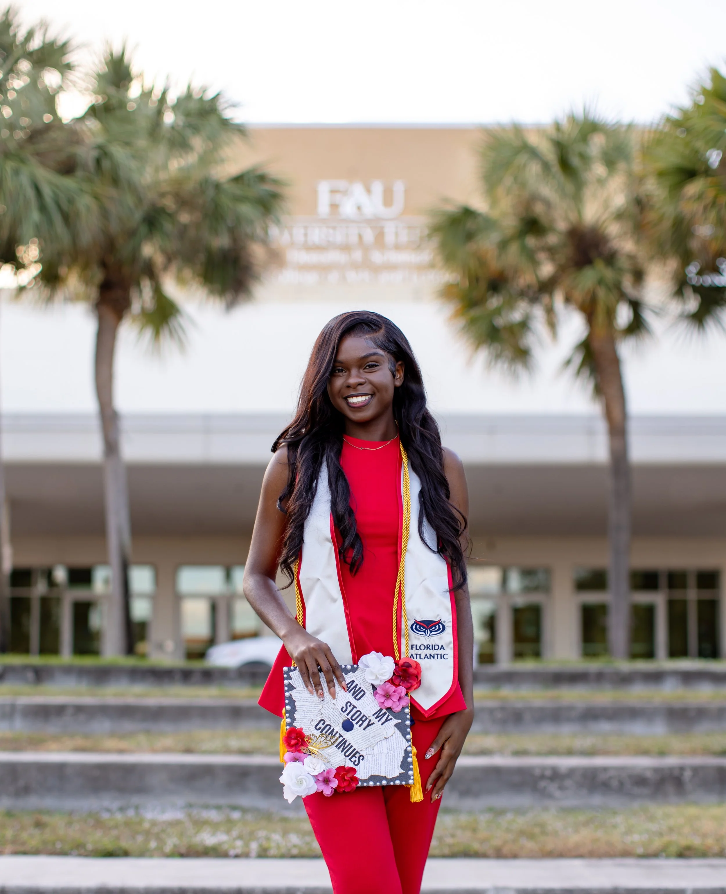 A young woman in a red dress and graduation stole holding a decorated diploma cover, standing outdoors in front of a building with palm trees.