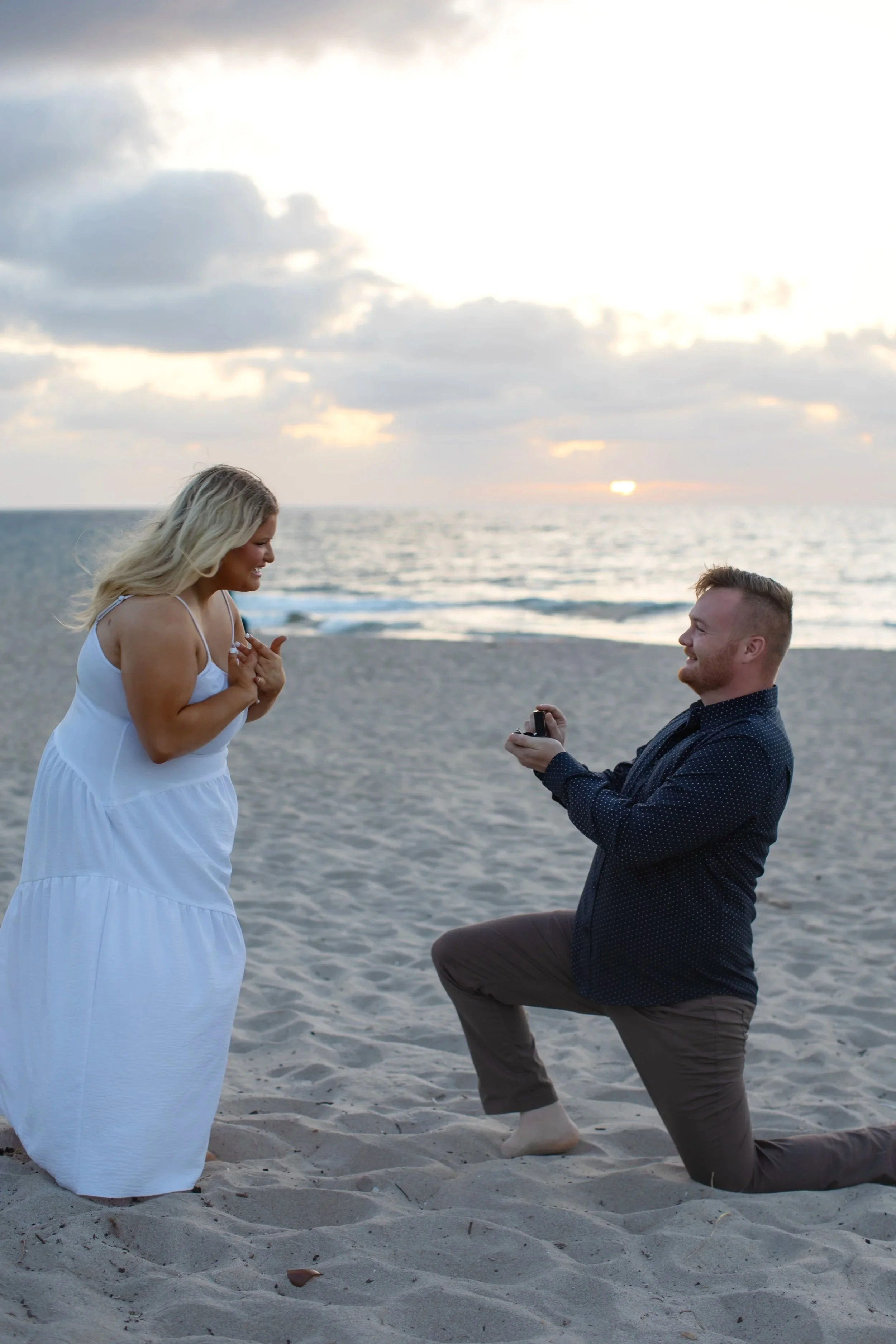 A man on one knee proposing to a woman on a beach at sunset, with the man holding a small ring box and the woman appearing surprised and happy.