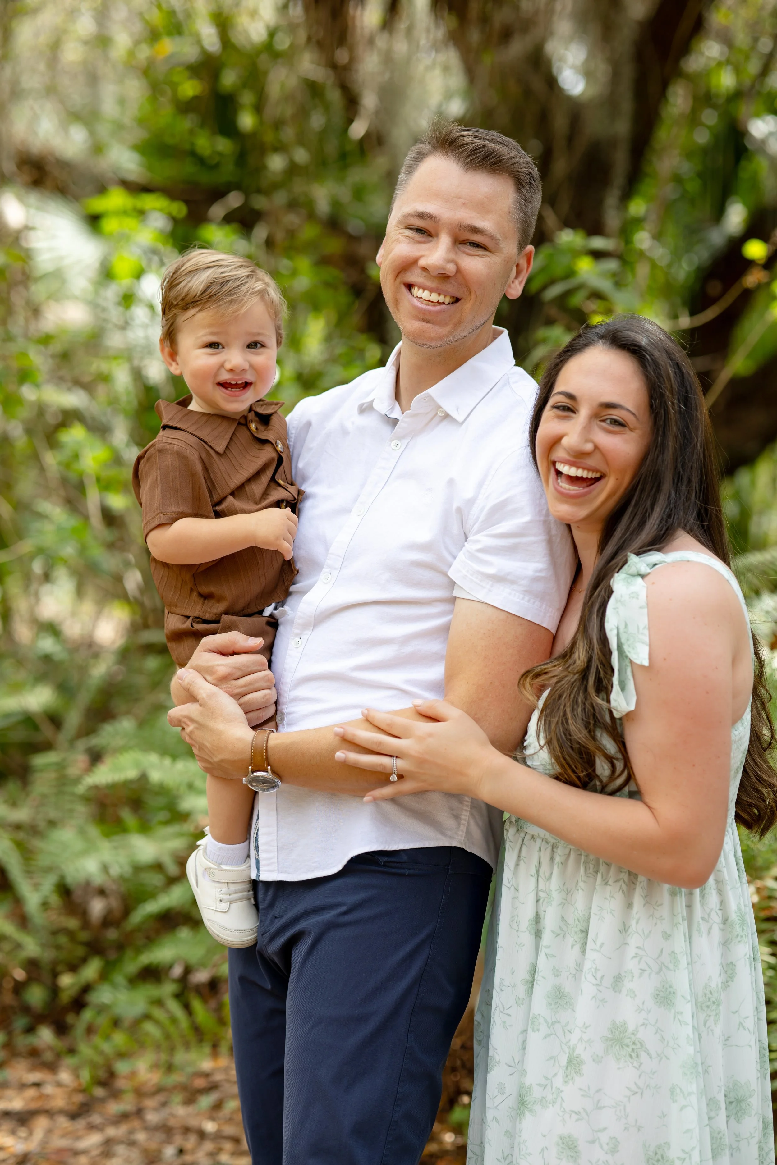 A joyful family of three outdoors, with a father holding a smiling young boy dressed in brown, and a smiling mother with long dark hair in a floral dress, all standing in a lush, green, wooded area.