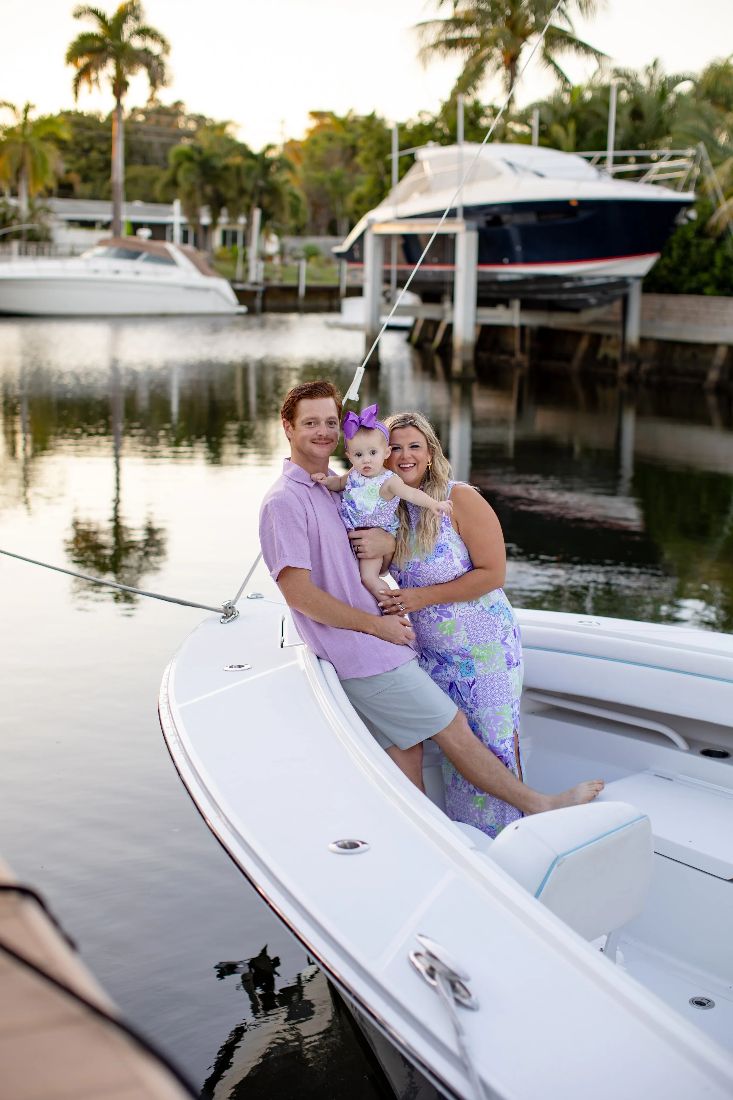 A family of three on a sailboat docked in a marina, with palm trees and boats in the background.