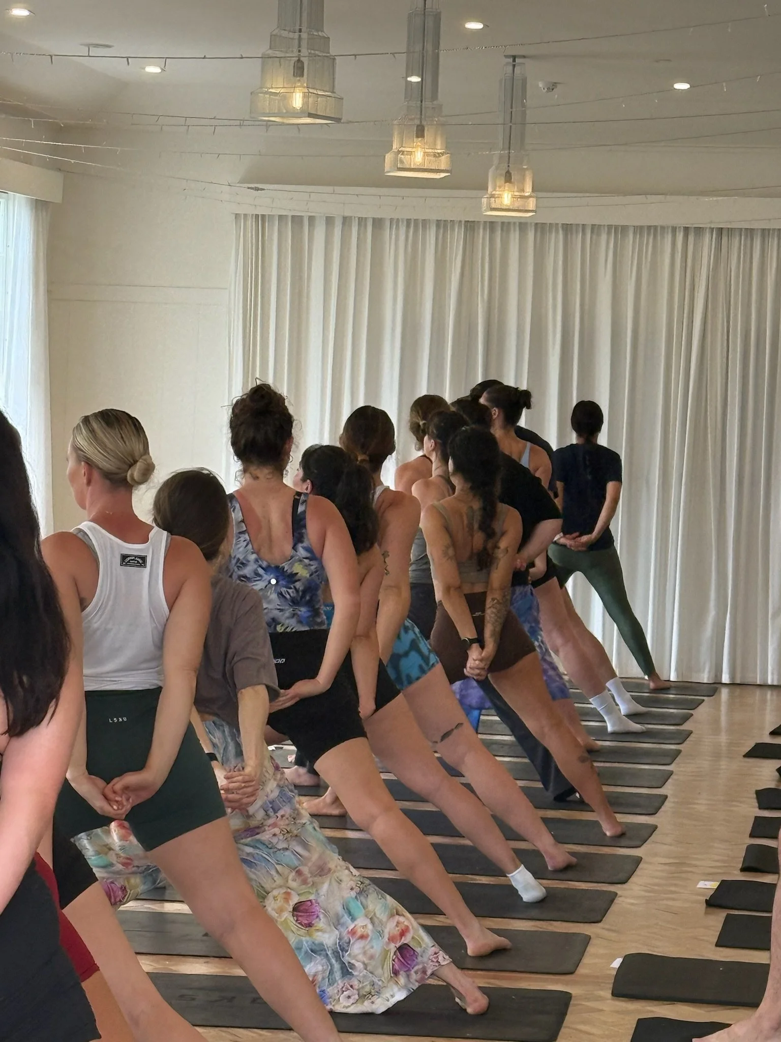 Group of women participating in a yoga class, practicing side stretches on yoga mats in a bright studio with white curtains.