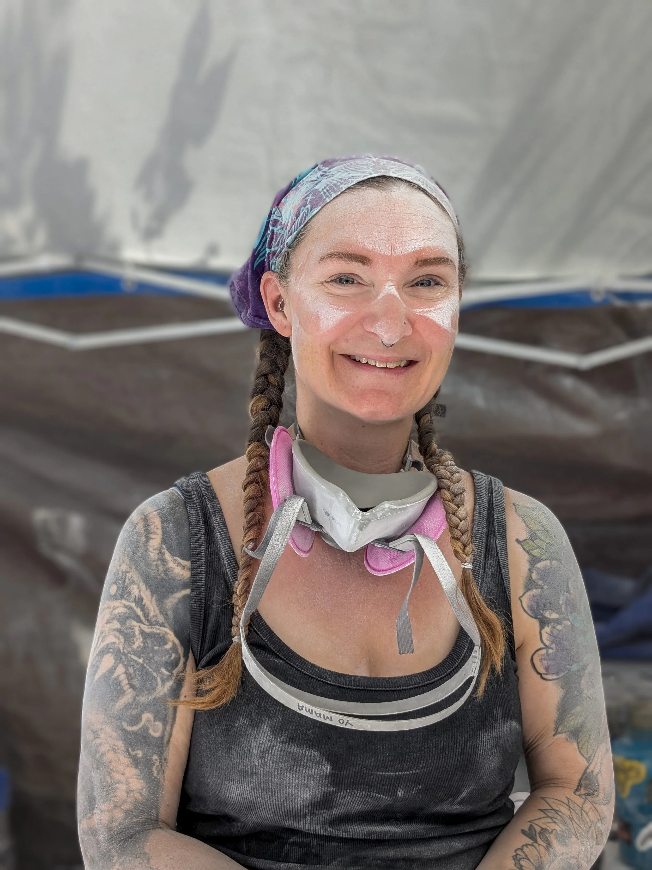 A woman with braids, tattoos, a bandana, and a respirator around her neck, smiling at the camera in a workshop or studio.