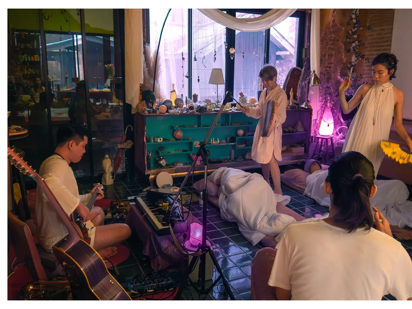 People perform a music and dance ritual indoors with candles and decorative objects around, some lying on the floor covered with white sheets, while others play guitar and keyboard or hold objects, creating a spiritual or artistic atmosphere.