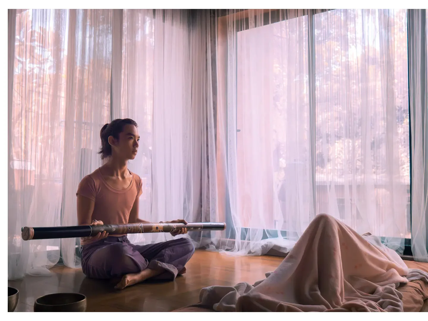 A young man sitting cross-legged on a wooden floor near large window with sheer curtains, holding a rainstick, with sunlight coming through pink-tinged trees outside.
