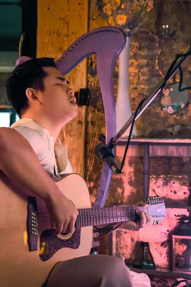 A young man playing an acoustic guitar and singing into a microphone in a cozy, warmly lit venue decorated with flowers and a harp in the background.