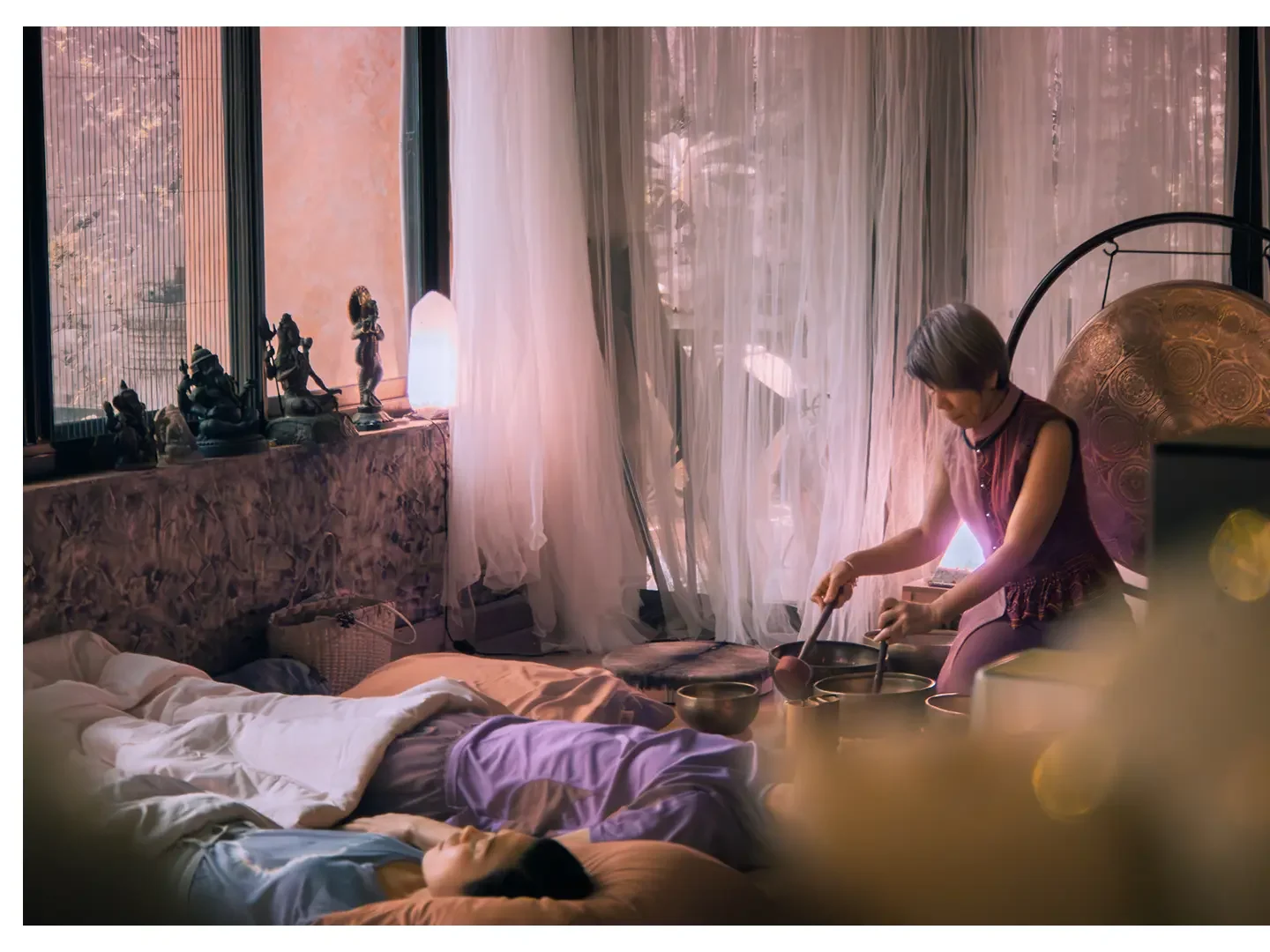 A woman with short hair is sitting on a floor, playing singing bowls, while a person is lying down on the bed, possibly asleep, in a room with large windows, sheer curtains, sunlight, and Asian-style ornaments and statues on the windowsill.
