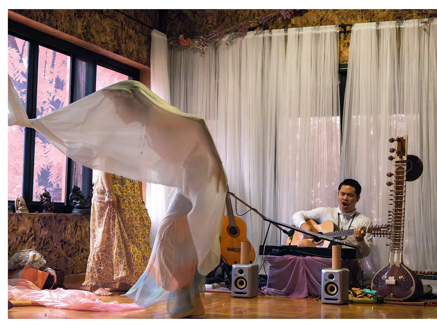Person dancing in front of a seated musician playing guitar, with musical instruments and speakers around, and a window with pink flowers outside.