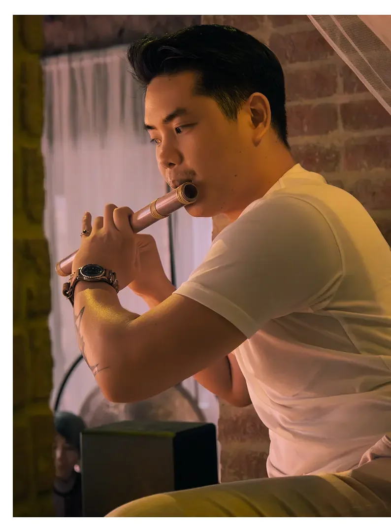 A young man with dark hair playing a flute indoors, with a brick wall and a window with white curtains in the background.