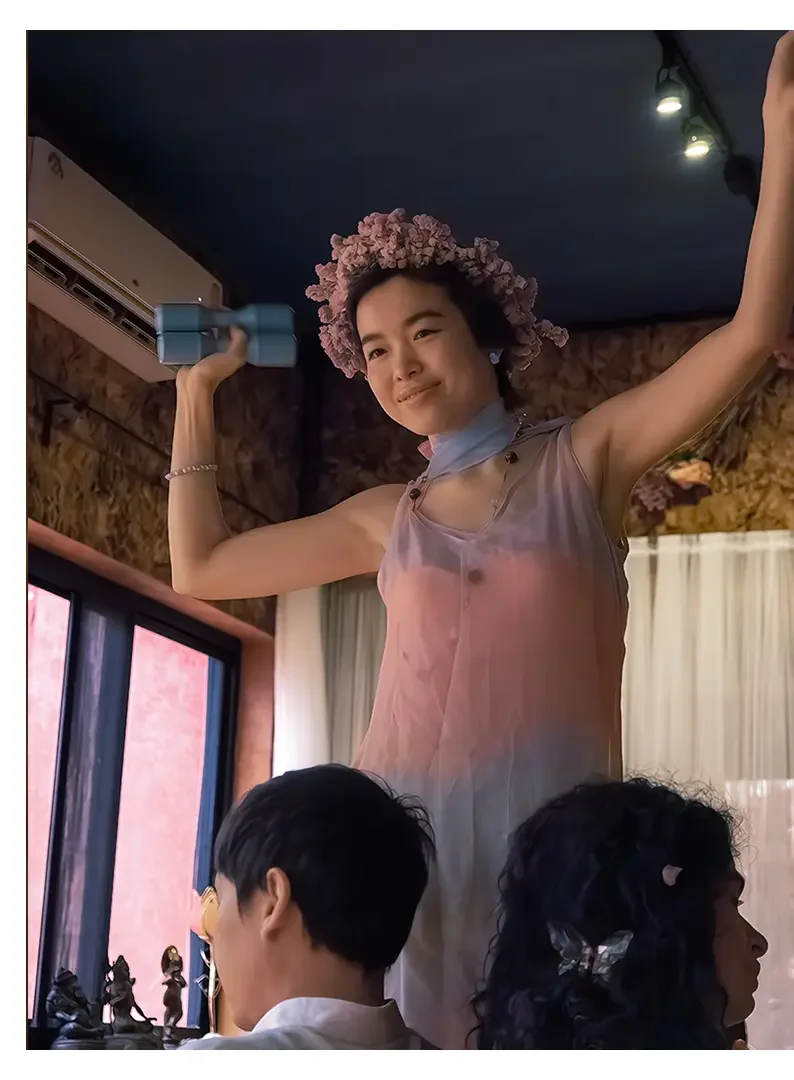 A young woman with a floral headband and light dress lifting a shaker while smiling, with two people sitting at a table in front of her, in a cozy indoor setting.