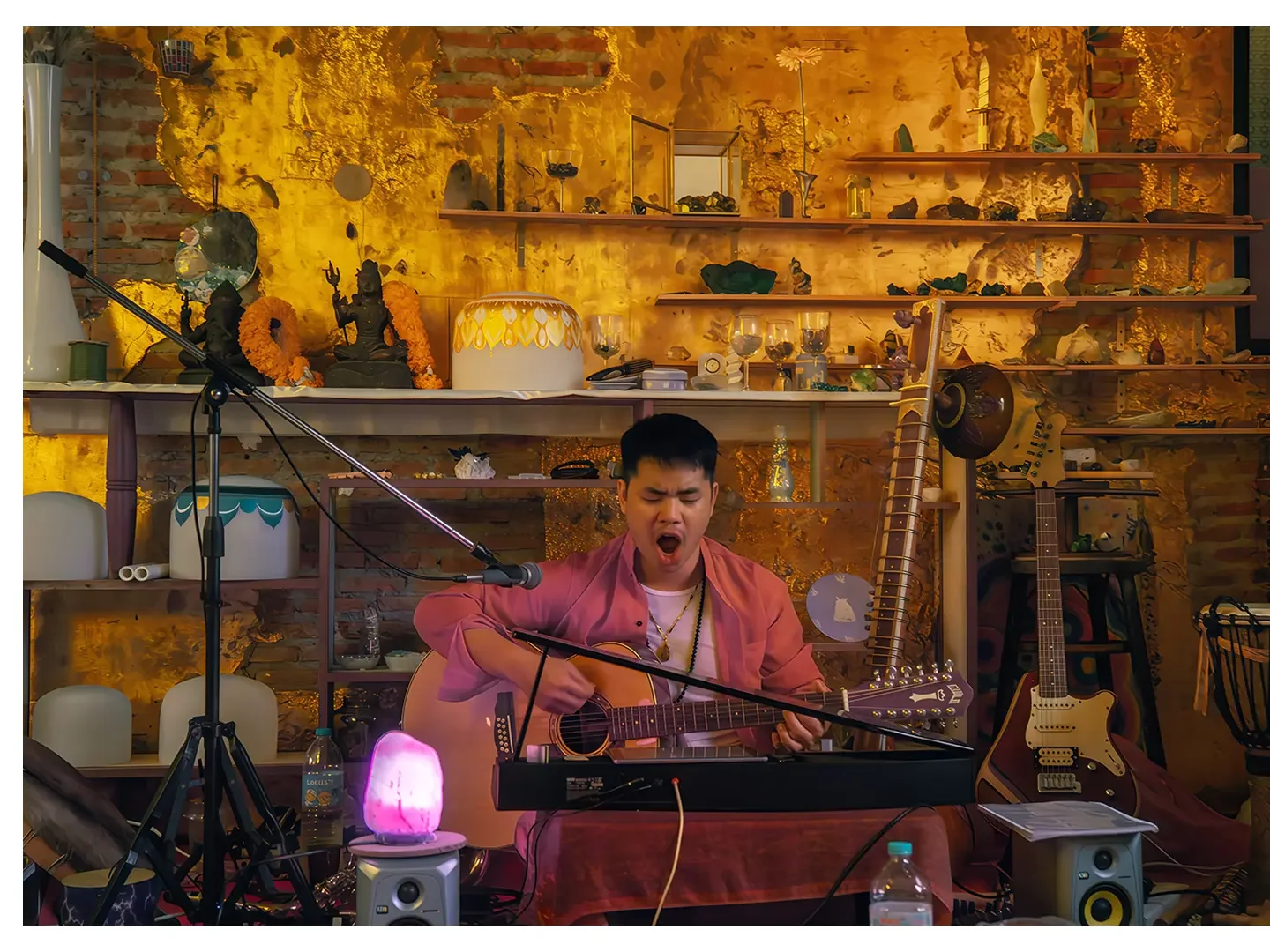 A man with short dark hair singing and playing an acoustic guitar and a keyboard in a cozy, decorated room with a guitar and electric guitar, shelves with ornaments, and a salt lamp.