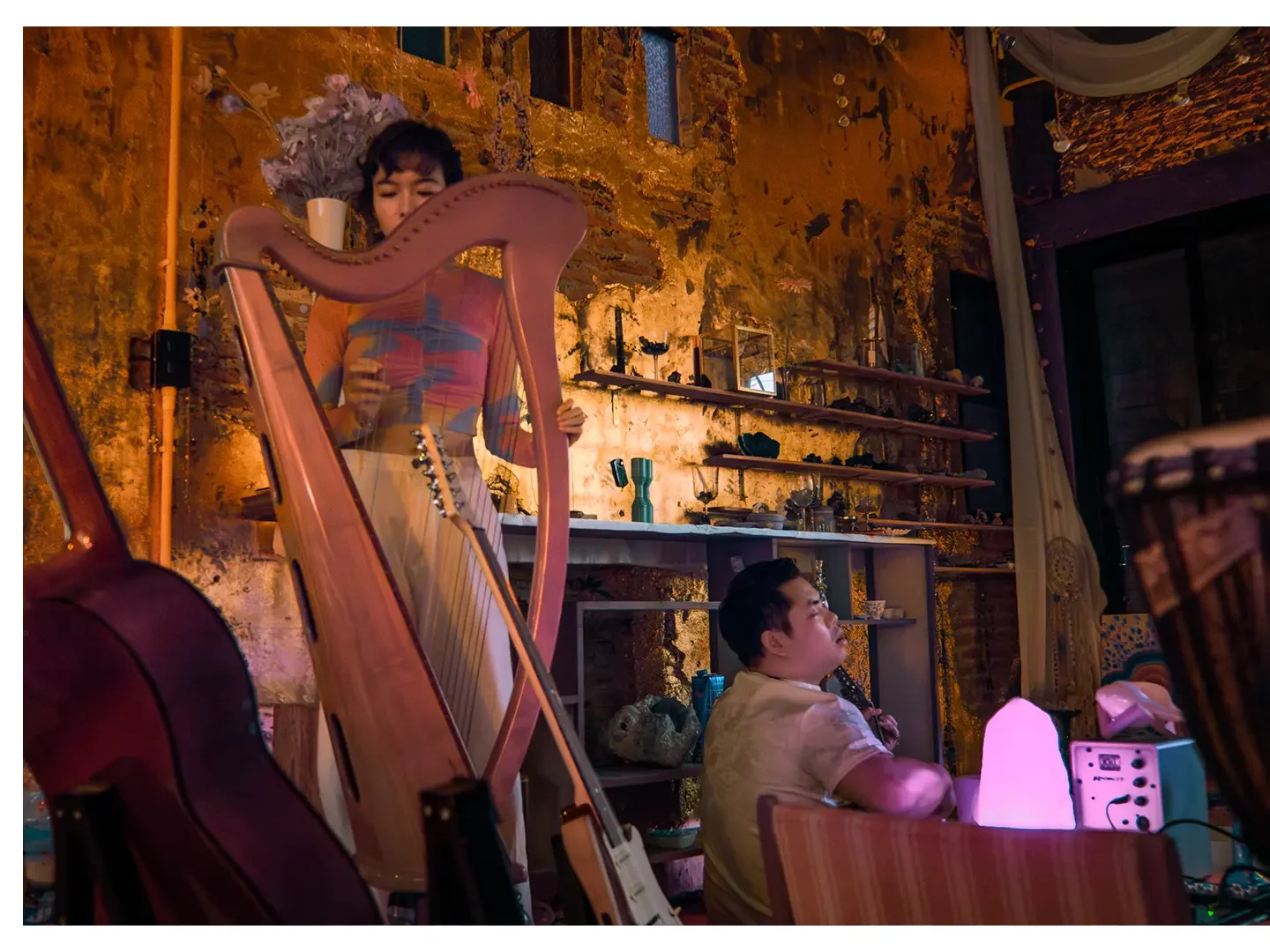 A woman playing a pink harp and a man sitting nearby singing into a microphone inside a warmly lit, decorated room.