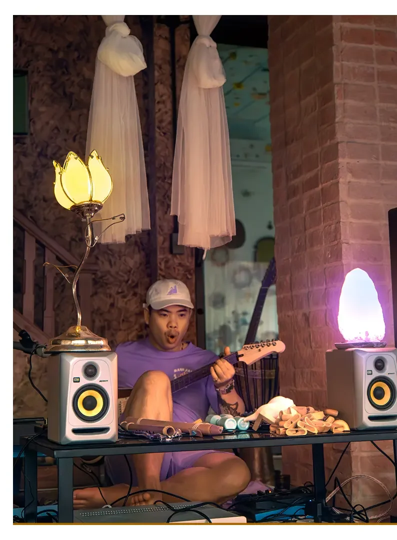 A young man wearing a gray cap and purple t-shirt, sitting cross-legged on the floor, playing a small guitar. He is surrounded by music equipment, props, and creative items on a black table, with decorative lamps and tulle hanging from the ceiling in