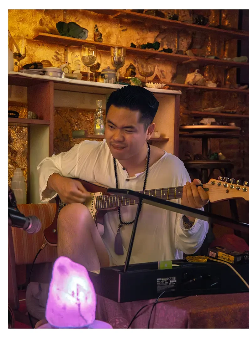 A man playing an electric guitar in a cozy, warmly lit room with shelves of decorative items and glassware in the background.