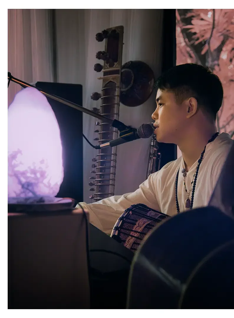 A young man with short dark hair playing a drum and singing into a microphone, with a purple-lit computer monitor in front of him, in a room decorated with musical instruments and a pink-flowered backdrop.