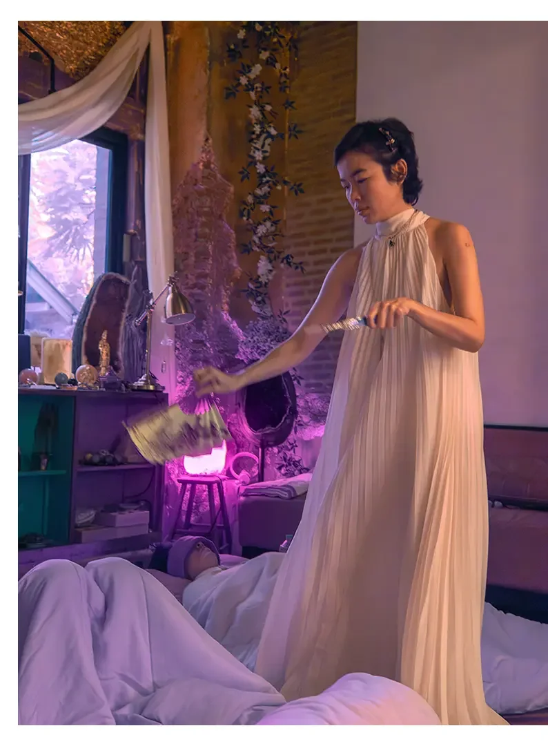 A woman wearing a long, pleated beige dress is standing in a cozy, dimly lit room with purple and pink lighting, holding a stack of money and reaching toward a person lying on the bed.