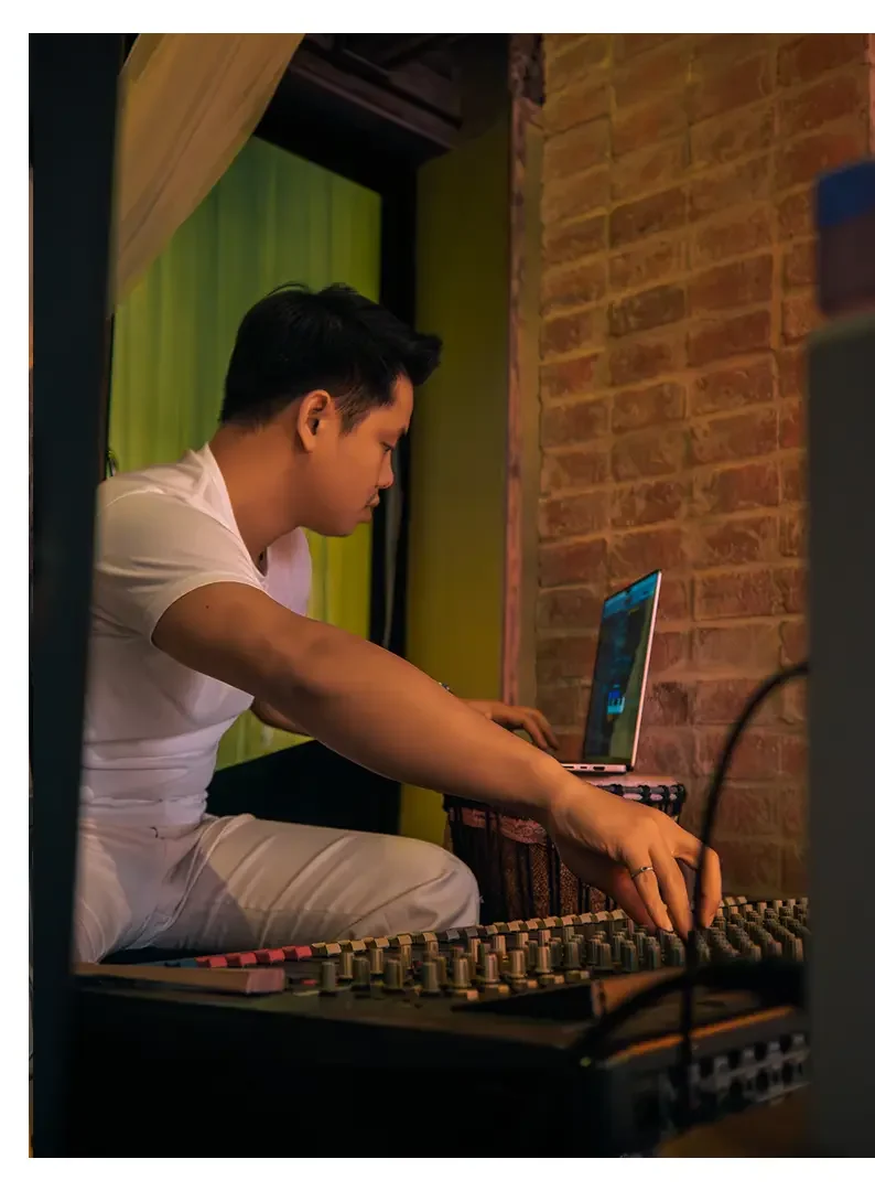 A person is sitting cross-legged near a brick wall, working with sound mixing equipment and a laptop.