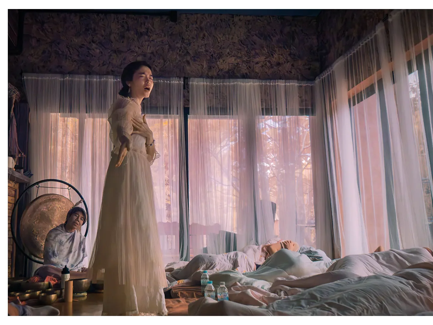 A woman in a long, light-colored dress appears to be singing with her hand on her chest in a bedroom with sheer curtains and sunlight coming through the windows. In the background, a person is lying on the bed, and another person is sitting on the fl