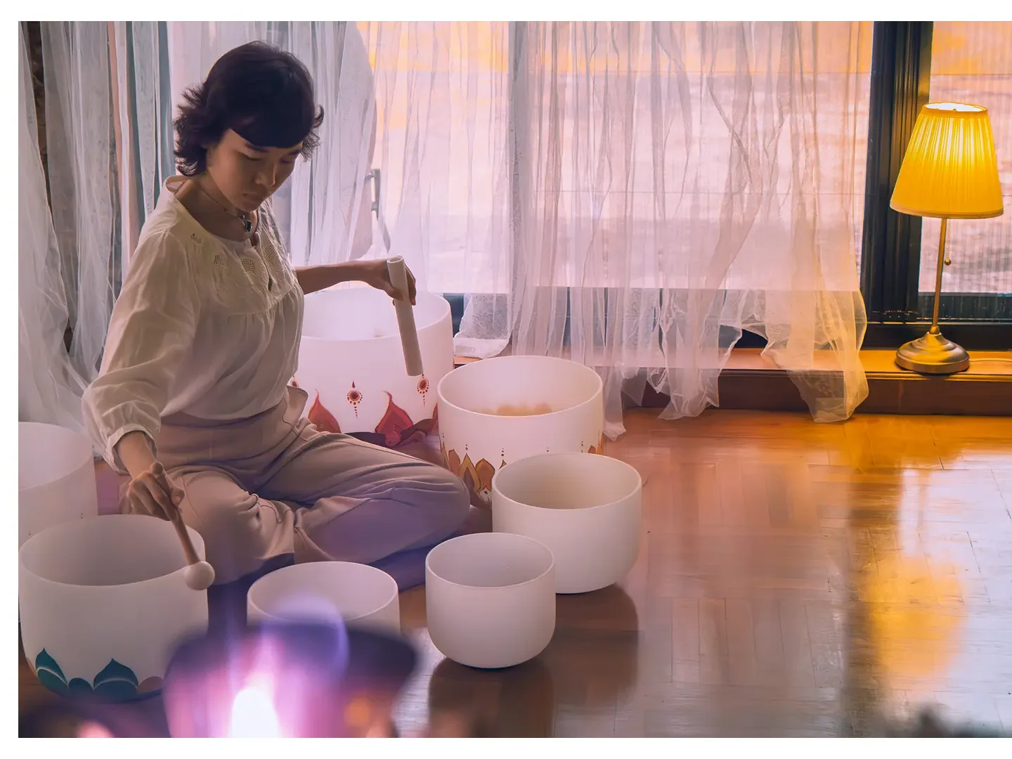 Woman sitting on the wooden floor playing 7 white singing bowls of different sizes in a room with sheer curtains and warm lighting.