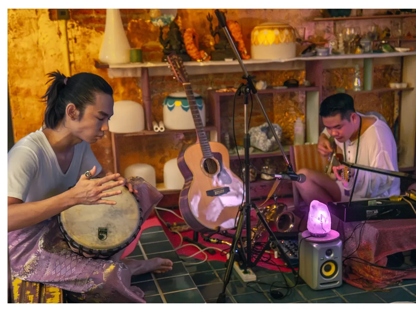 Two musicians playing instruments in a cozy, decorated indoor space with guitars, a salt lamp, and various decor items on shelves.