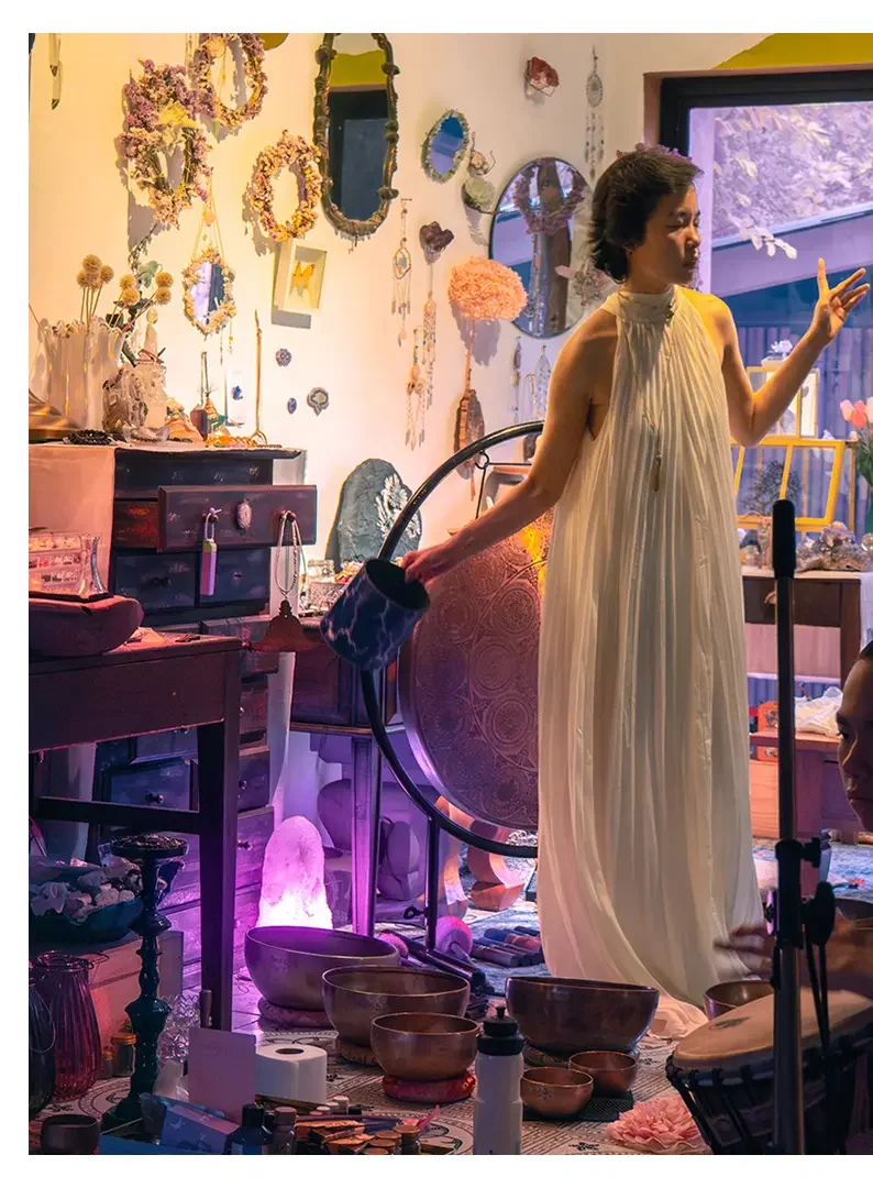 A woman in a white, pleated dress making a peace sign with her fingers inside a colorful, decorated room filled with mirrors, jewelry, crystals, bowls, and various decorative items.