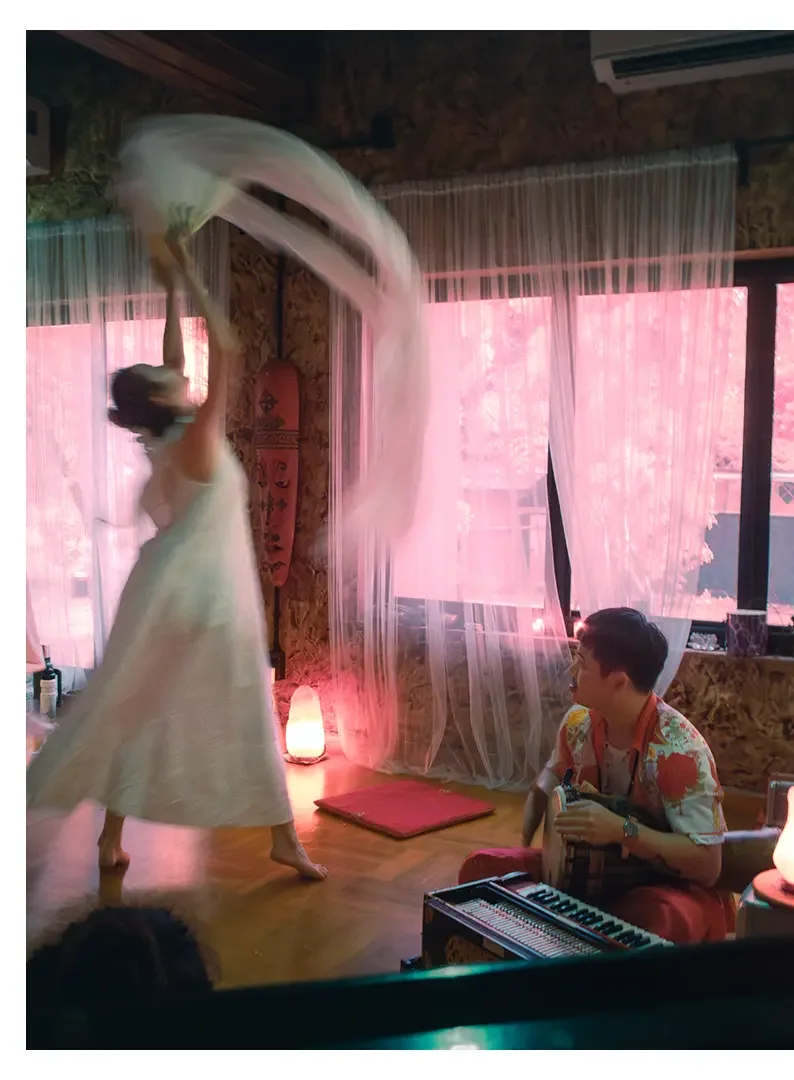 A woman dancing with a fan in a room with pink curtains and a wooden floor. A man sitting nearby, wearing a colorful shirt, holding a guitar. There is a small keyboard and a glowing lamp nearby.
