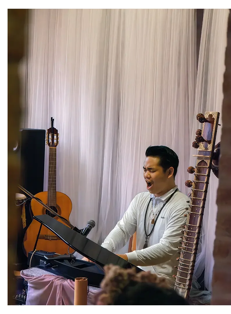 A man singing passionately while playing the keyboard, with a guitar in the background and traditional musical instruments nearby, in a room with white curtains.