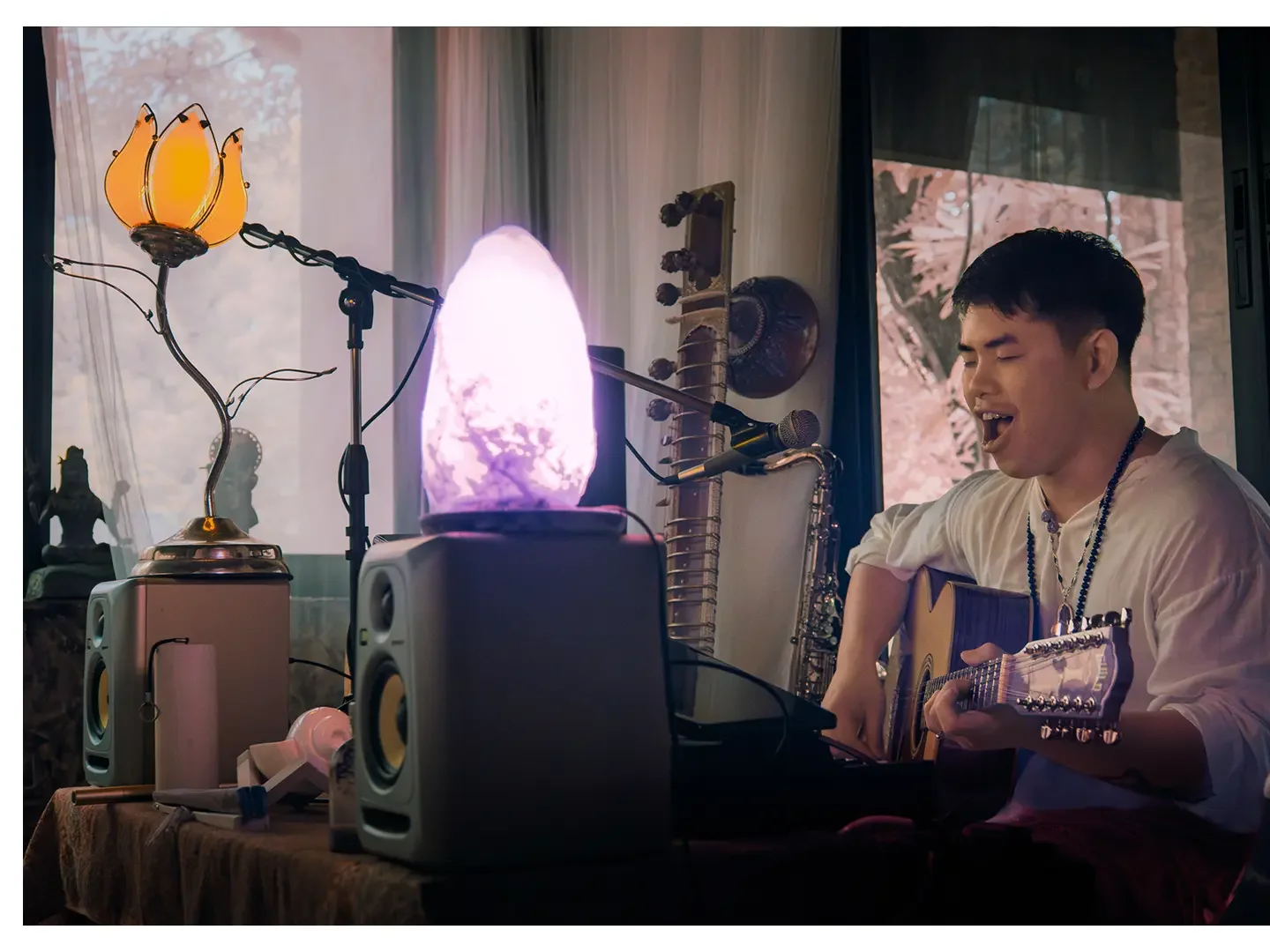 A young man sitting indoors, singing passionately and playing an acoustic guitar, with various musical equipment including speakers, a microphone, and a Himalayan salt lamp emitting purple light, in a cozy room decorated with curtains and art.