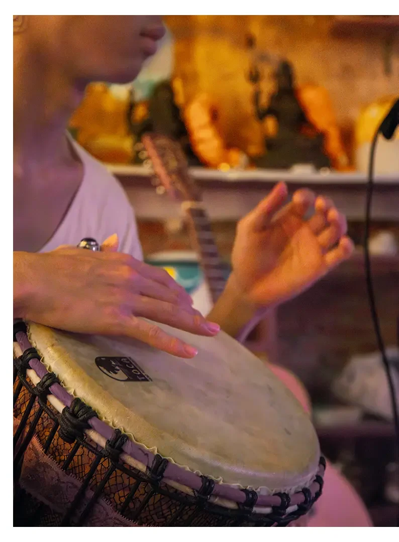 Close-up of a person playing a djembe drum with hands on the drumhead, in a setting with wooden and musical instrument background.