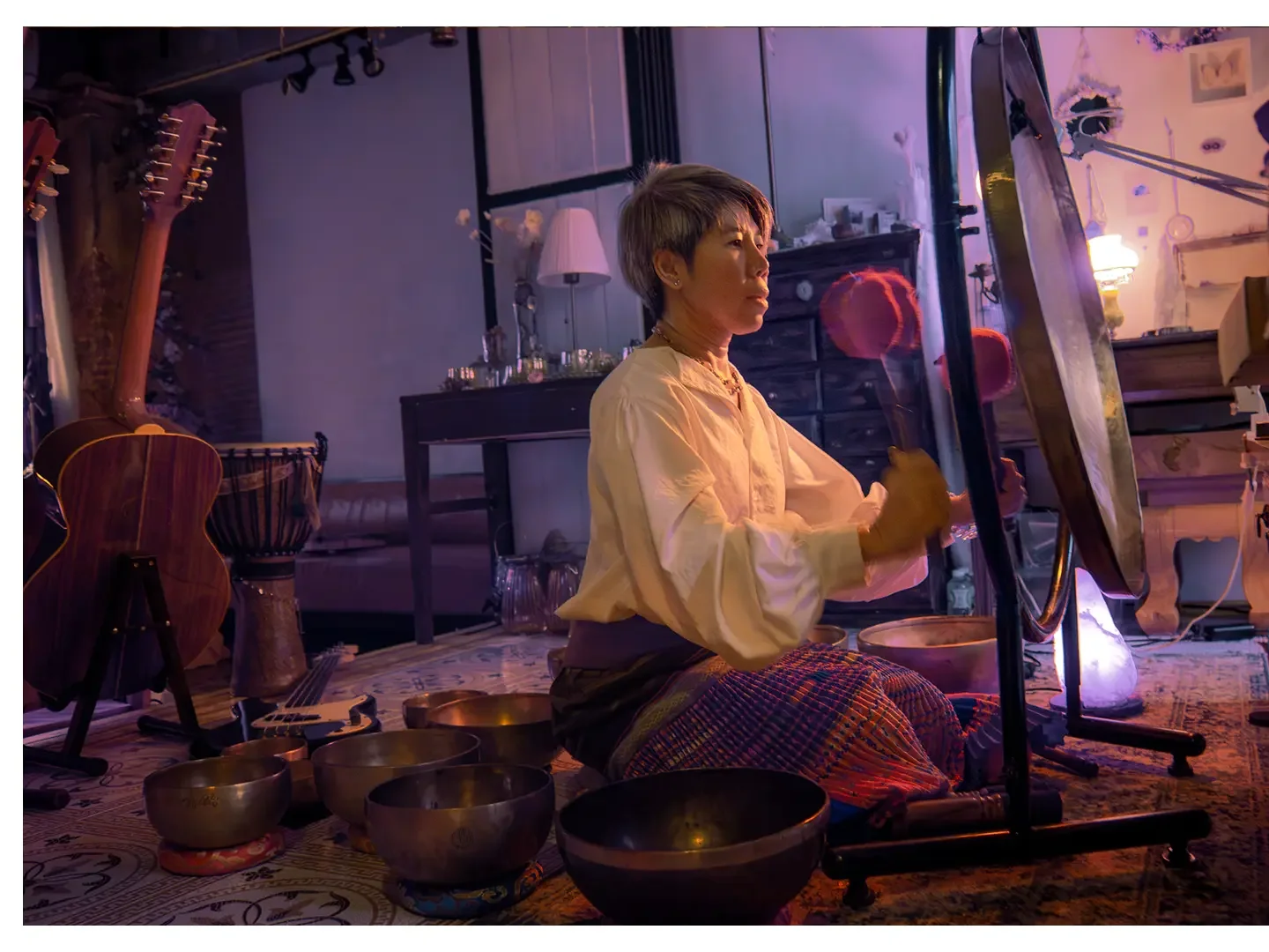 A woman with short hair sits cross-legged on the floor, surrounded by singing bowls, in a dimly lit room with musical instruments and furniture, as she appears to be performing a spiritual ritual.