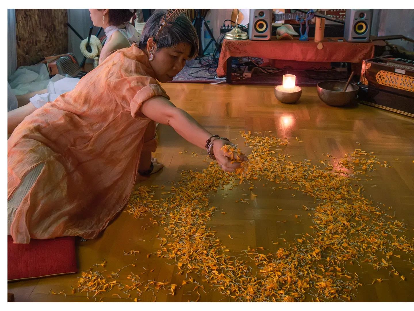 Woman in an orange dress kneeling on a wooden floor, spreading yellow flower petals, with a candle, bowls, and music equipment in the background.