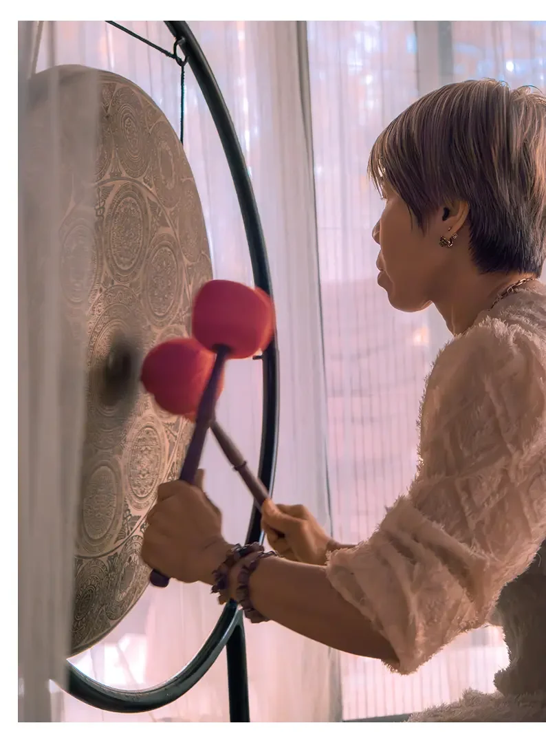 A woman with short hair playing a gong with a pink mallet in a room with sheer curtains.