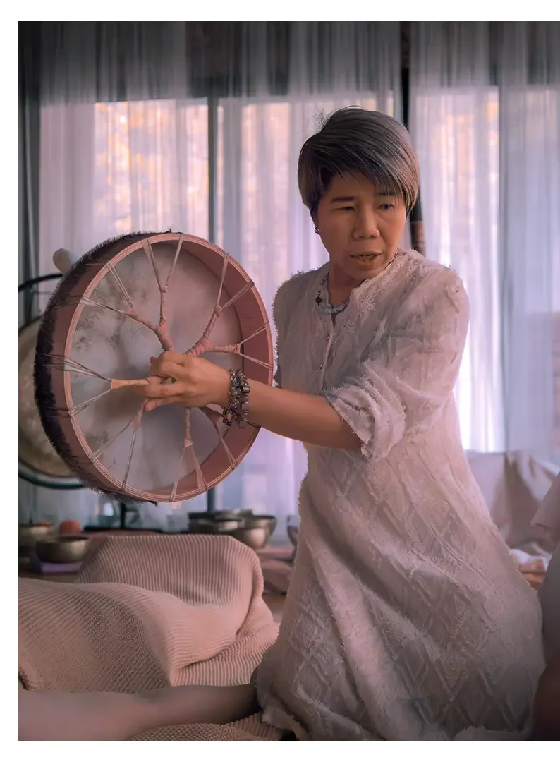 A woman with short gray hair wearing a pale pink dress, holding a drum with a wooden frame and animal hide, in a room with soft pink and purple lighting, curtains, and a sofa.