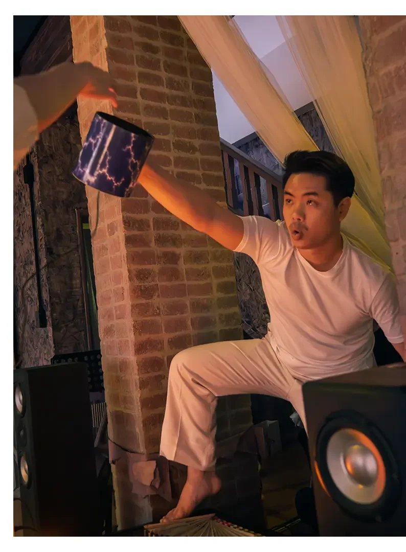 A young man wearing a white t-shirt and beige pants seated with one leg up on a piece of furniture, reaching out with his arm to make a sound, in a room with brick walls and sound equipment.