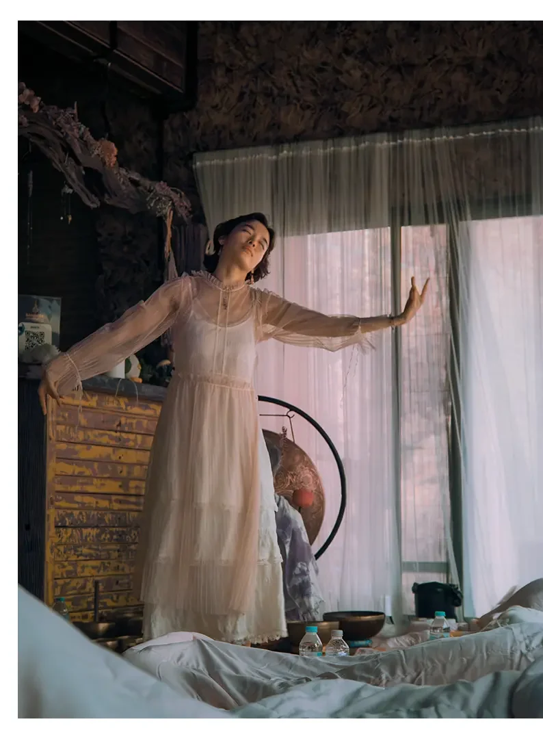 A woman in a sheer, beige dress poses with her arms outstretched inside a cozy room with large windows covered by sheer curtains and a wooden ceiling